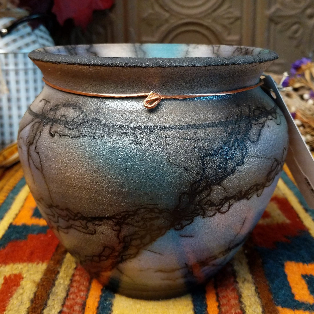 This beautiful Mottled Blue Horsehair Pottery by Roger Calhoun features a rounded ceramic vessel with mottled blue and turquoise tones, black horsehair patterns, a flared rim, and decorative copper wire with a turquoise-colored bead.