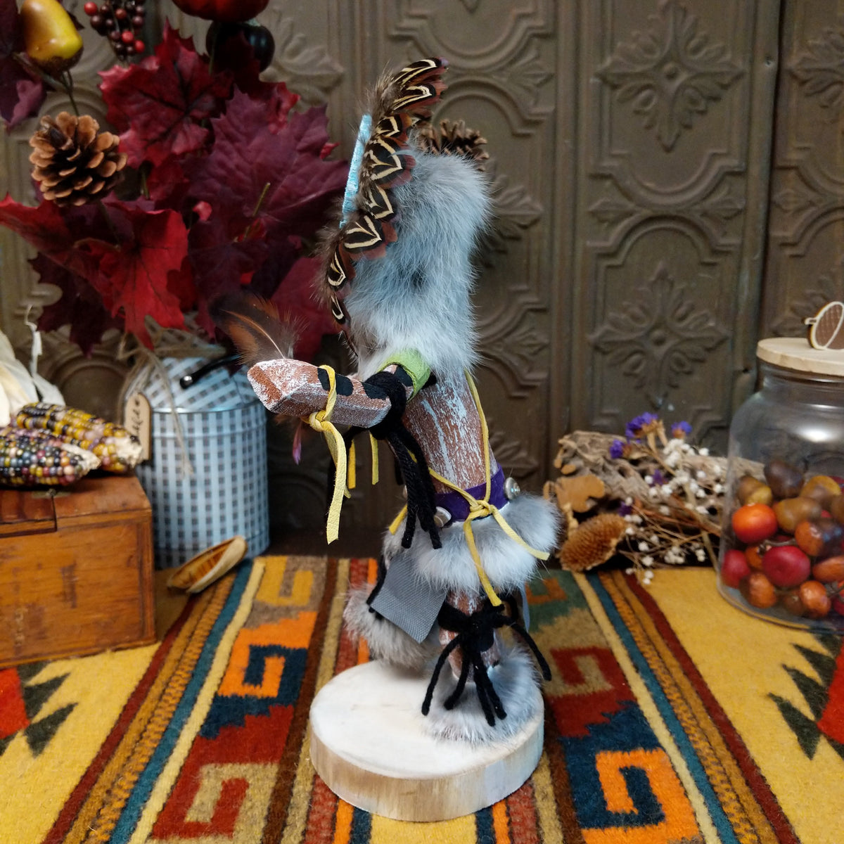 Side view of the Navajo Sunface Kachina doll with turquoise-colored face, feather headdress, outstretched arms, and blue fabric clothing.