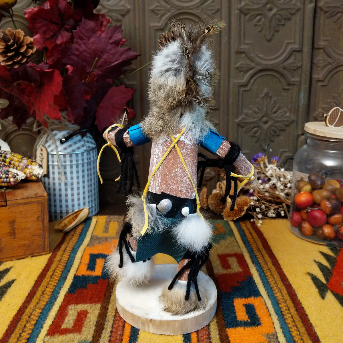 Back view of the Navajo Protection Dancer Kachina doll with gray fur headdress, black painted facial markings, blue sleeves, feather accents, and raised foot.