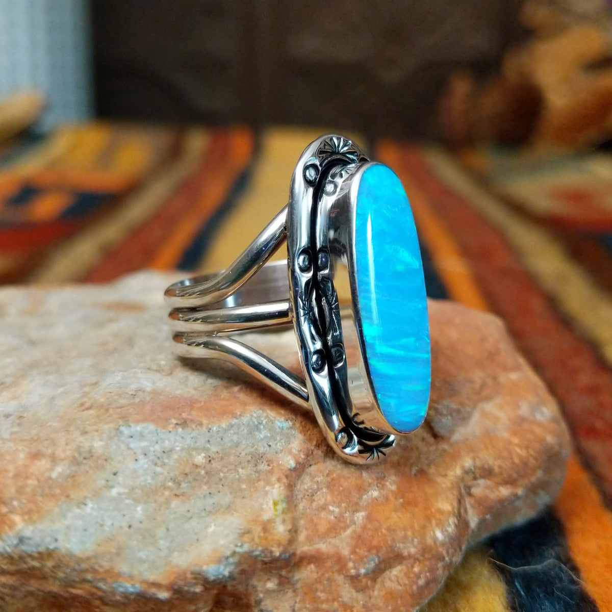 Blue Manmade Opal Ring by Terry Wood featuring an elongated oval blue opal with iridescent tones, decorative stamped sterling silver bezel, raised silver beads, and split-shank band; side view.