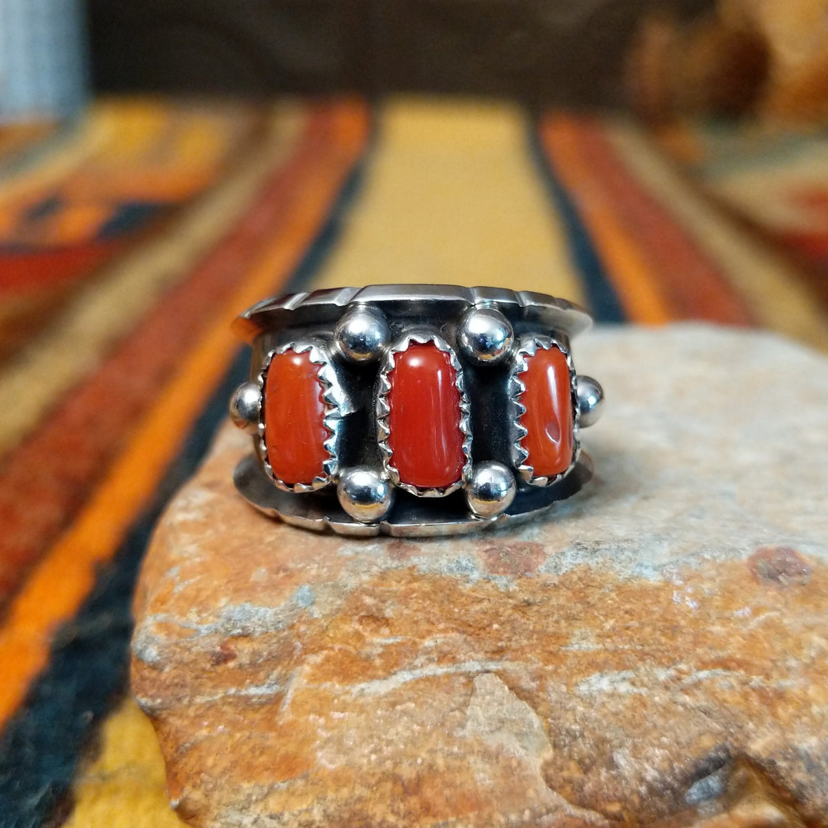 This striking Coral Trio Ring by Reed Charley features three rectangular red-orange coral stones in sawtooth silver bezels, accented by polished silver beads and a wide oxidized silver band.