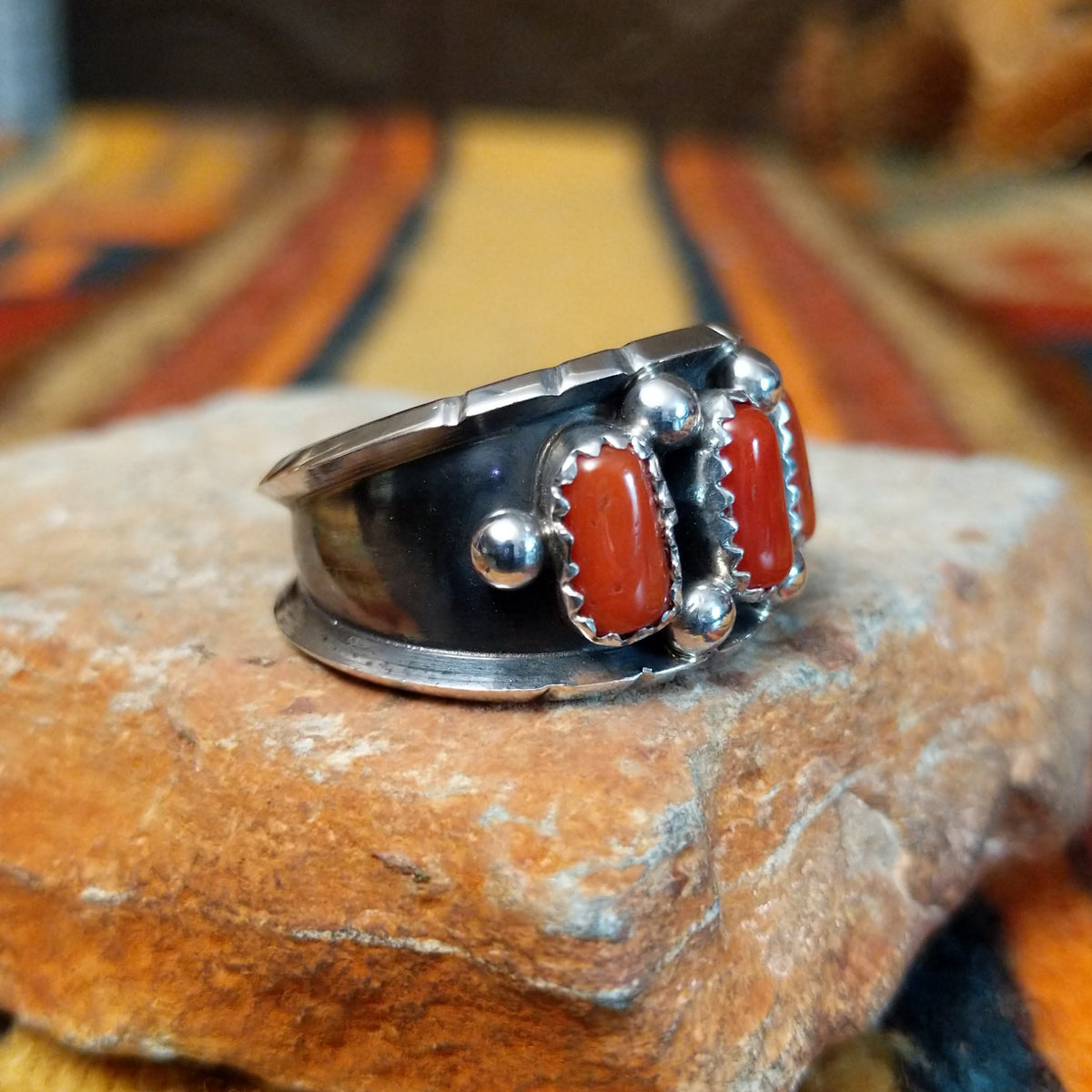 This striking Coral Trio Ring by Reed Charley features three rectangular red-orange coral stones in sawtooth silver bezels, accented by polished silver beads and a wide oxidized silver band; side view.