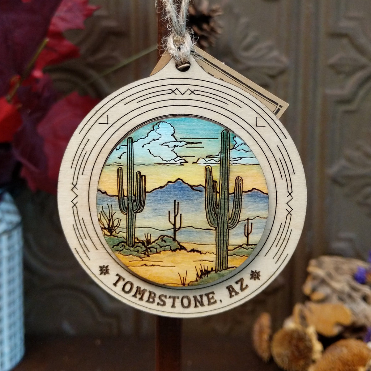 Desert Saguaro Sunrise Wood Ornament featuring tall green cacti, distant blue mountains, colorful sunrise sky, engraved circular wooden frame, “Tombstone, AZ” lettering, and natural twine hanging loop.