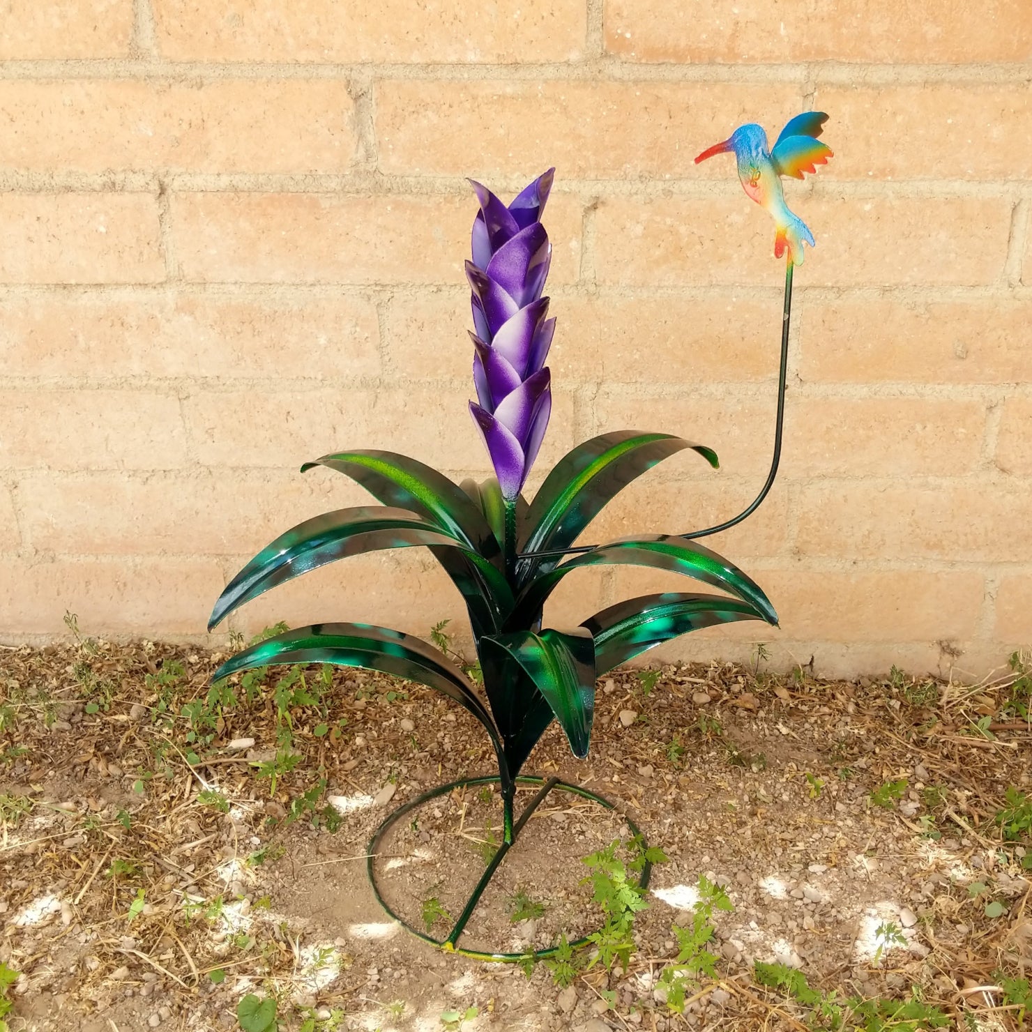 A large purple metal flower with green leaves and a multicolored hummingbird perched above.