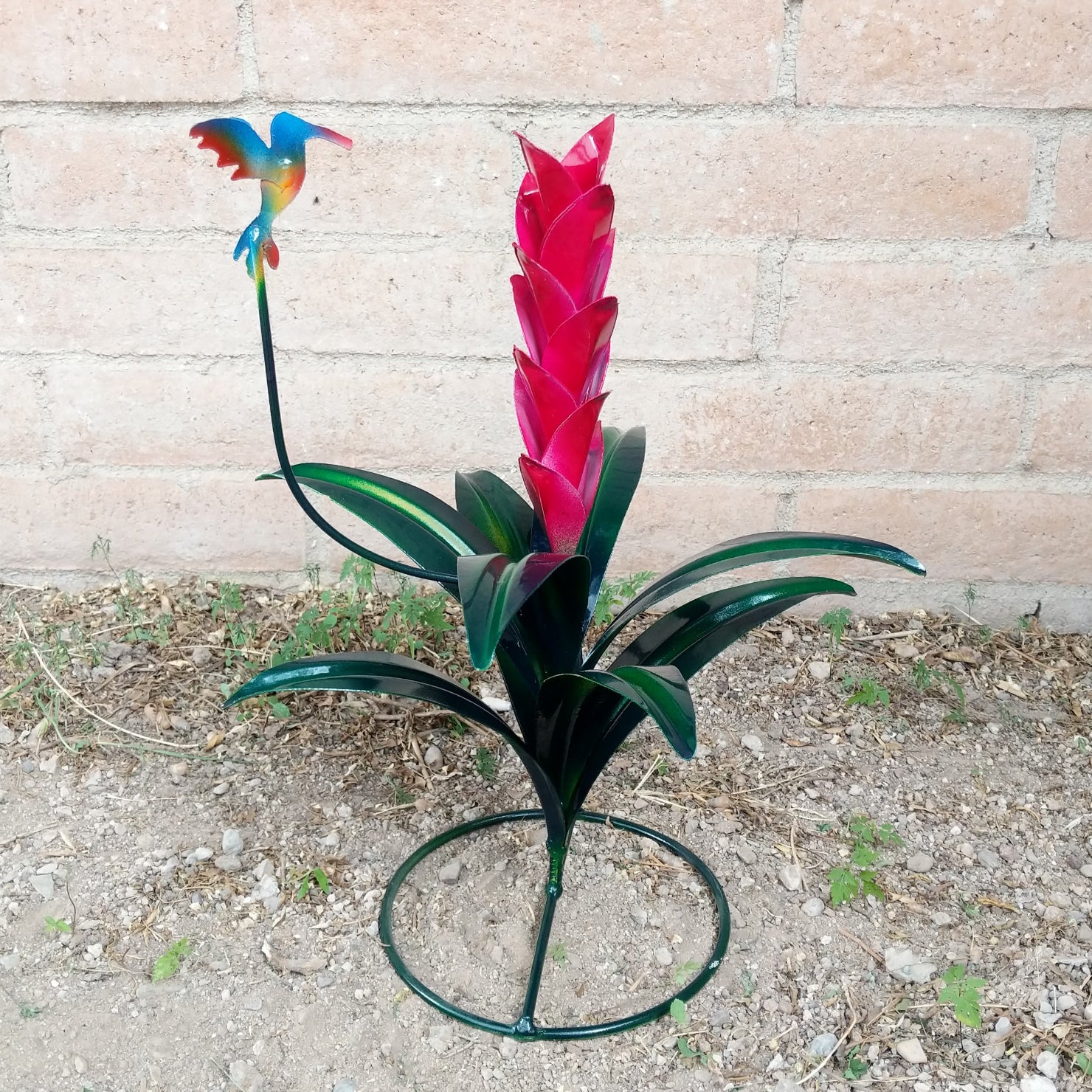 A small magenta metal flower with green leaves and a multicolored hummingbird perched above.