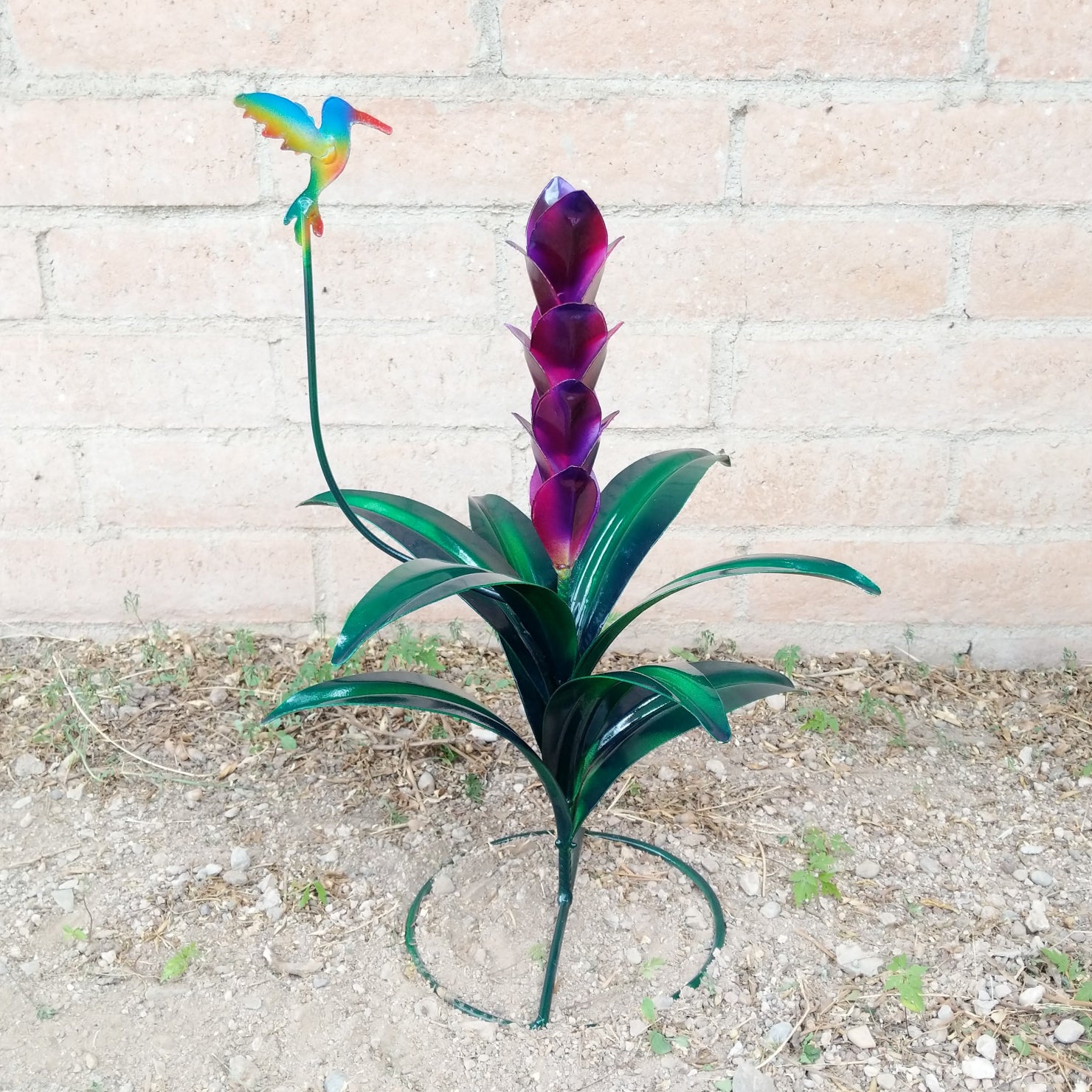 A small purple metal flower with green leaves and a multicolored hummingbird perched above.