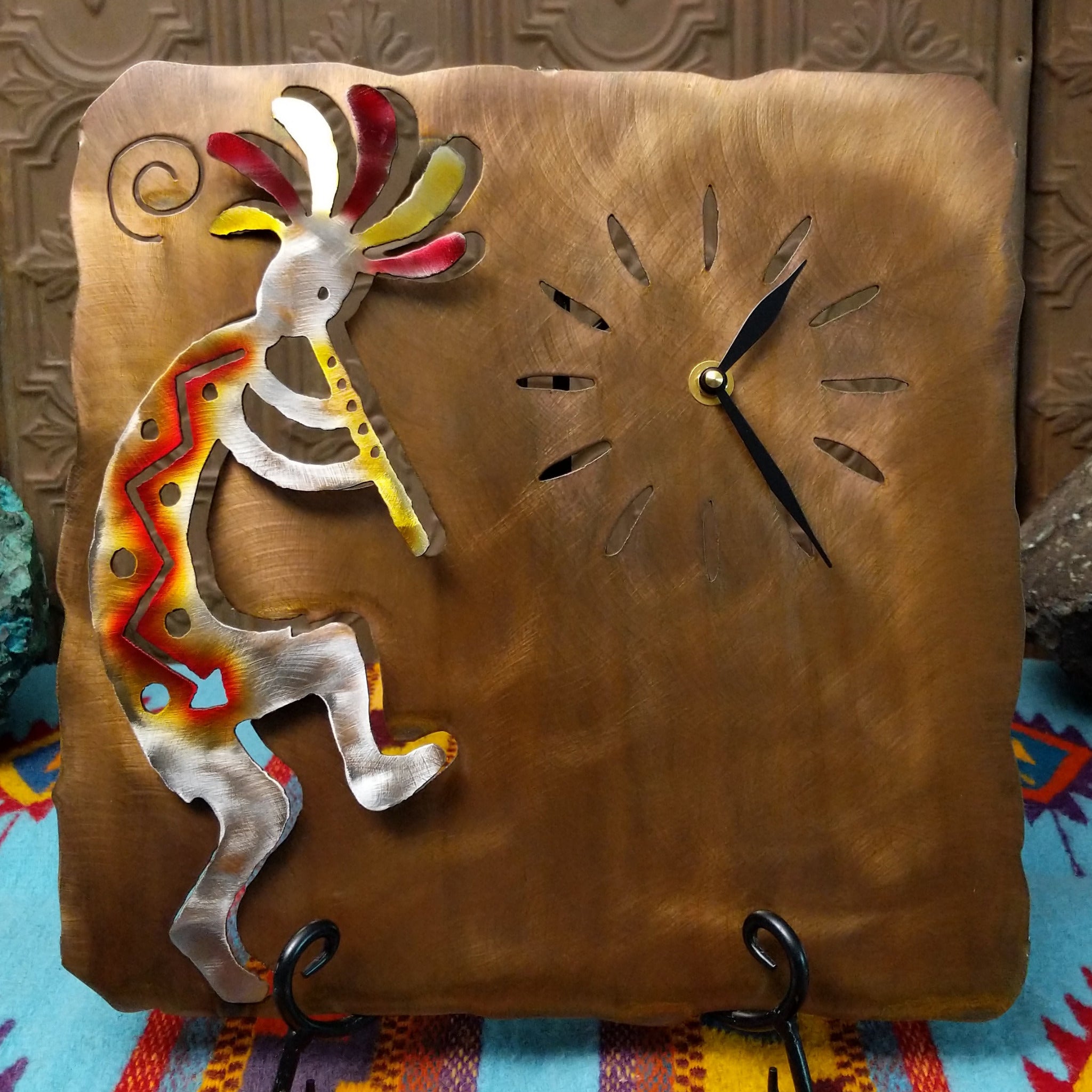 This Crooks Design metal clock features a silver-toned flute-playing Kokopelli with red and yellow Sunset Swirl accents, cutout hour markers, black clock hands, and a warm textured metal background.