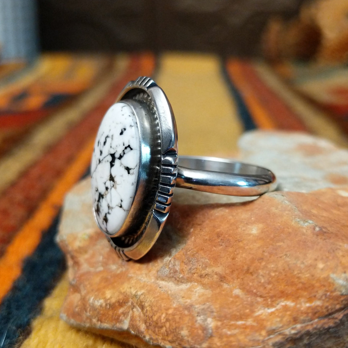 This distinctive White Buffalo Ring by Alfred Martinez features a large oval white stone with black matrix patterns, surrounded by a silver rope border and decorative stamped silver setting; side view.
