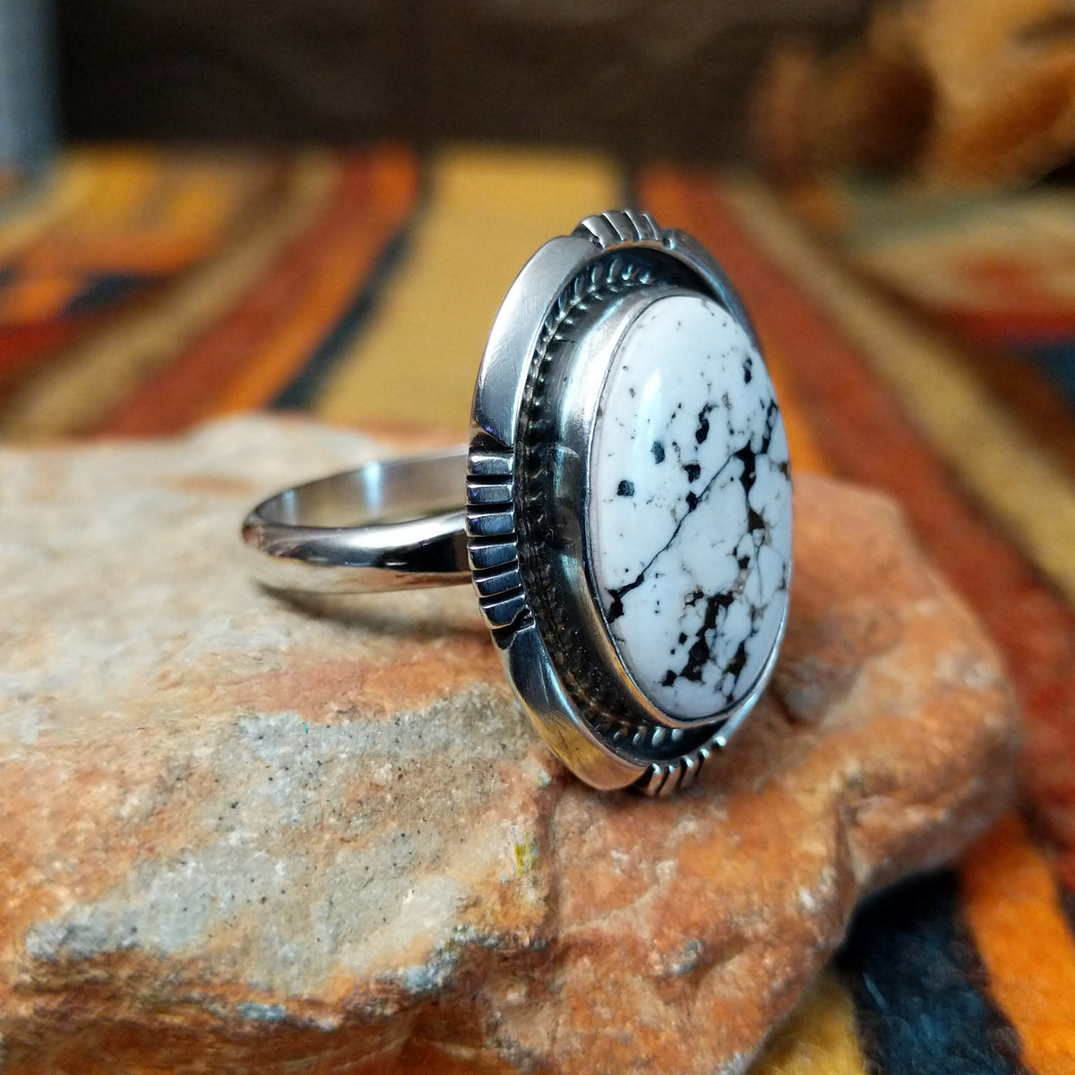This distinctive White Buffalo Ring by Alfred Martinez features a large oval white stone with black matrix patterns, surrounded by a silver rope border and decorative stamped silver setting; side view.