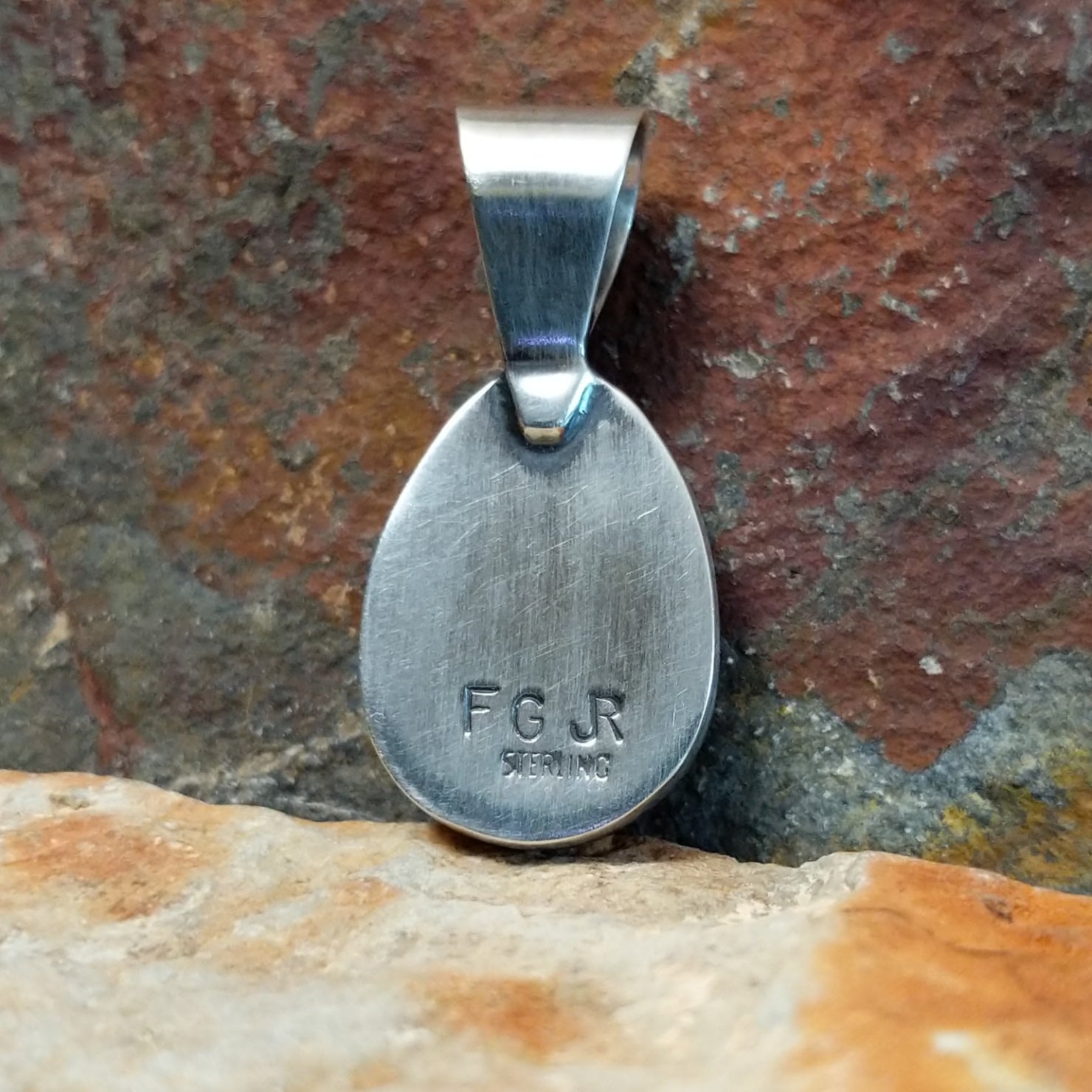 The stamped view of this turquoise pendant displays a maker’s mark etched into the silver base near the smooth bezel.