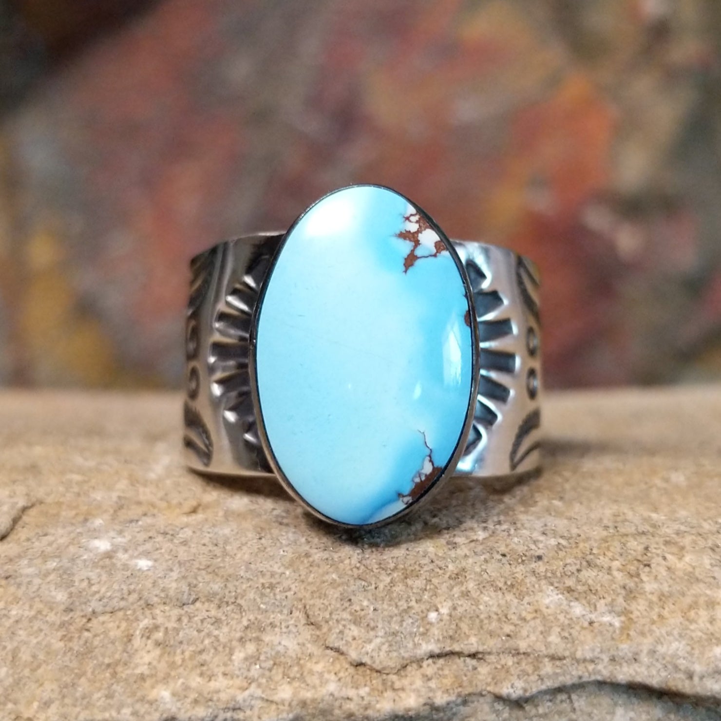 A large, oval Golden Hills turquoise ring by Bernyse Chavez & Lowell Draper, featuring a sky-blue stone with brown matrix veining, set in a wide sterling silver band with a symmetrical sun-ray pattern. The ring rests on a textured sandstone surface with a warm, earthy red background.