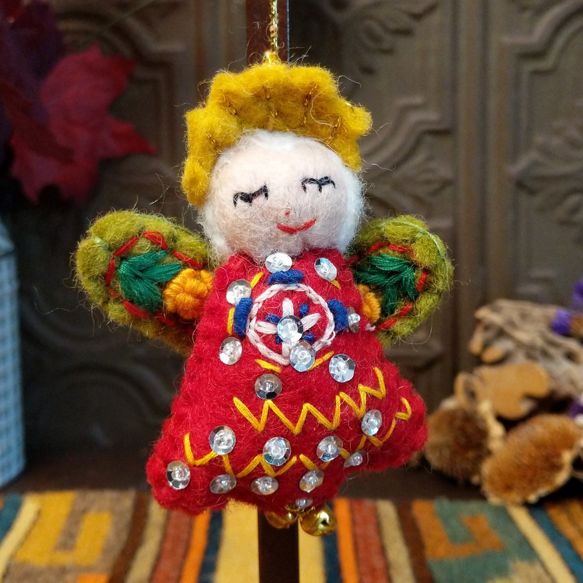 This whimsical Green-Winged Angel Wool Ornament features golden yellow hair, a smiling white face, embroidered green wings, a red dress with colorful stitching and silver sequins, dangling gold bells, and a gold hanging cord.