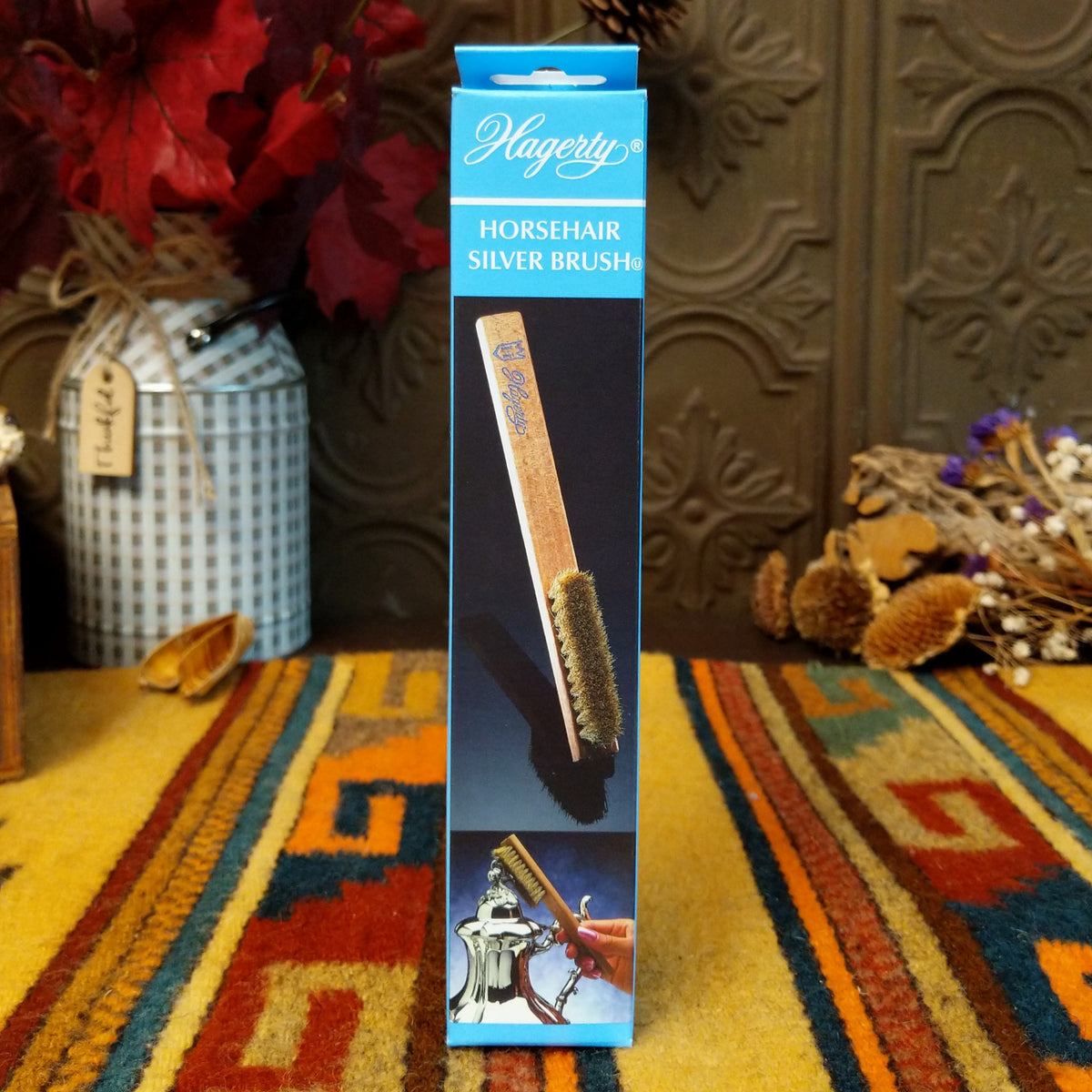 A boxed horsehair silver brush standing upright on a patterned rug in a styled indoor setting with decorative dried flowers and a dark carved wall behind it.