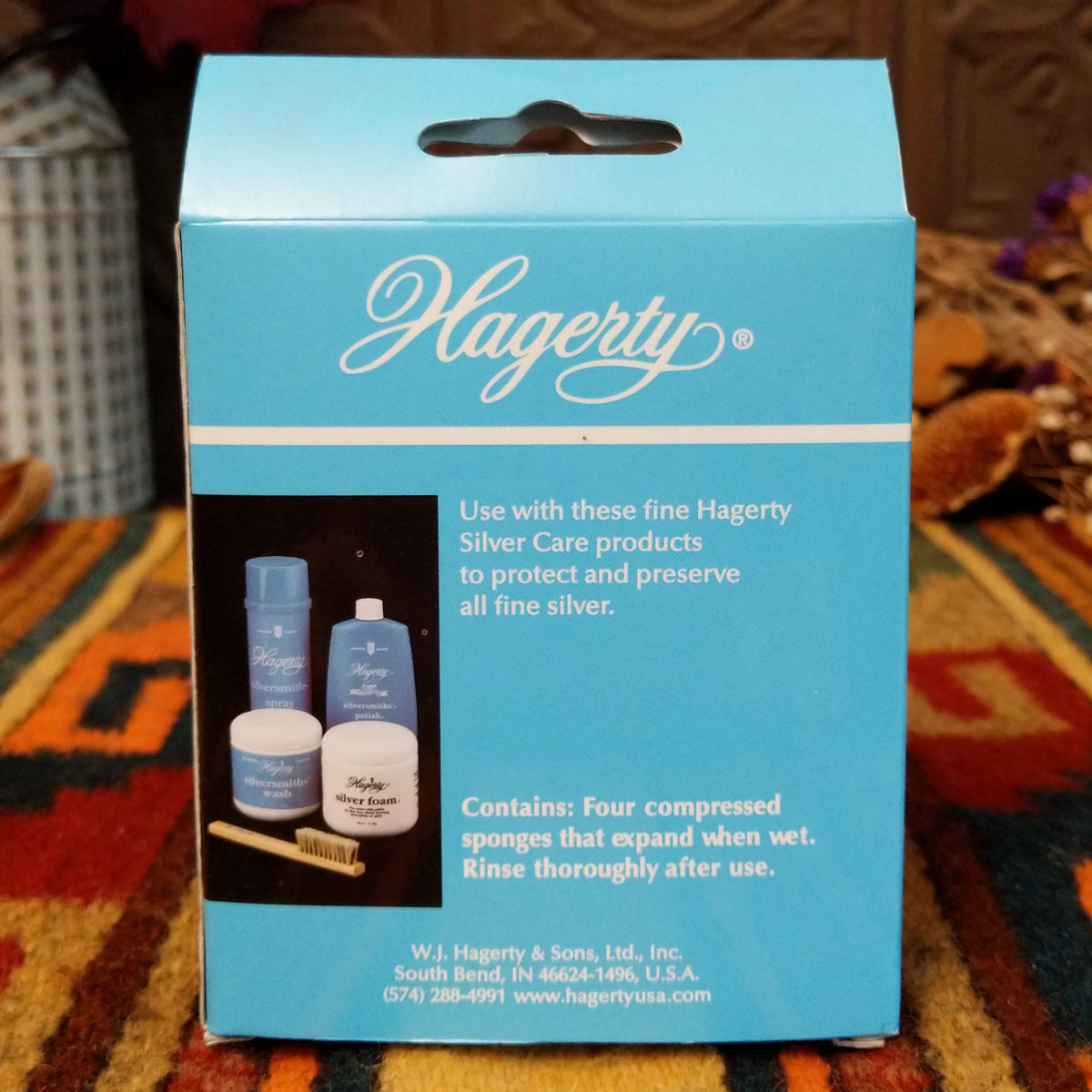 A light blue Hagerty product box standing on a patterned orange and brown rug, with a dark wood carved backdrop and dried floral decor in the background.