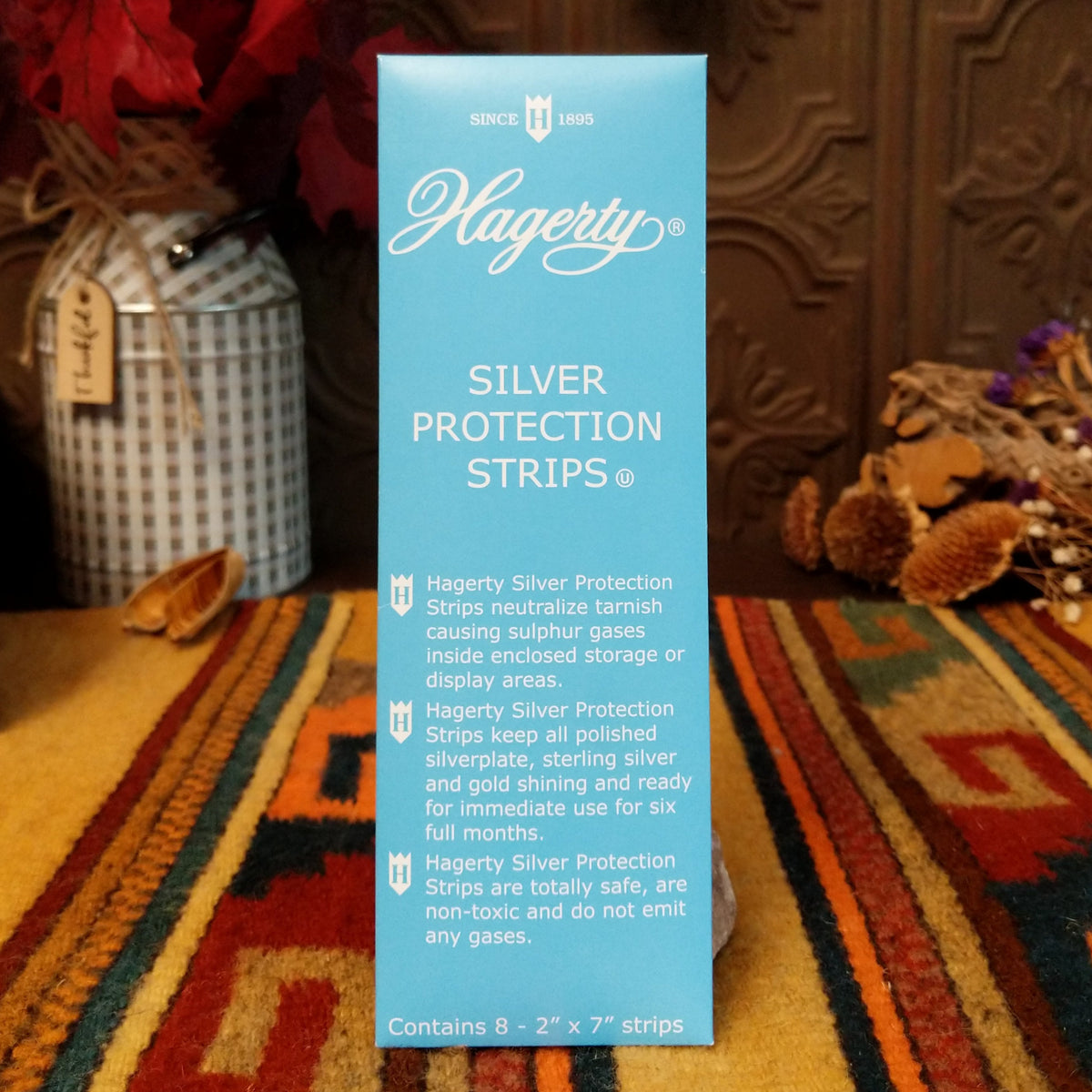 A box of Hagerty Silver Protection Strips stands upright on a patterned rug in a styled indoor setting with decorative dried flowers and a dark carved wooden background.
