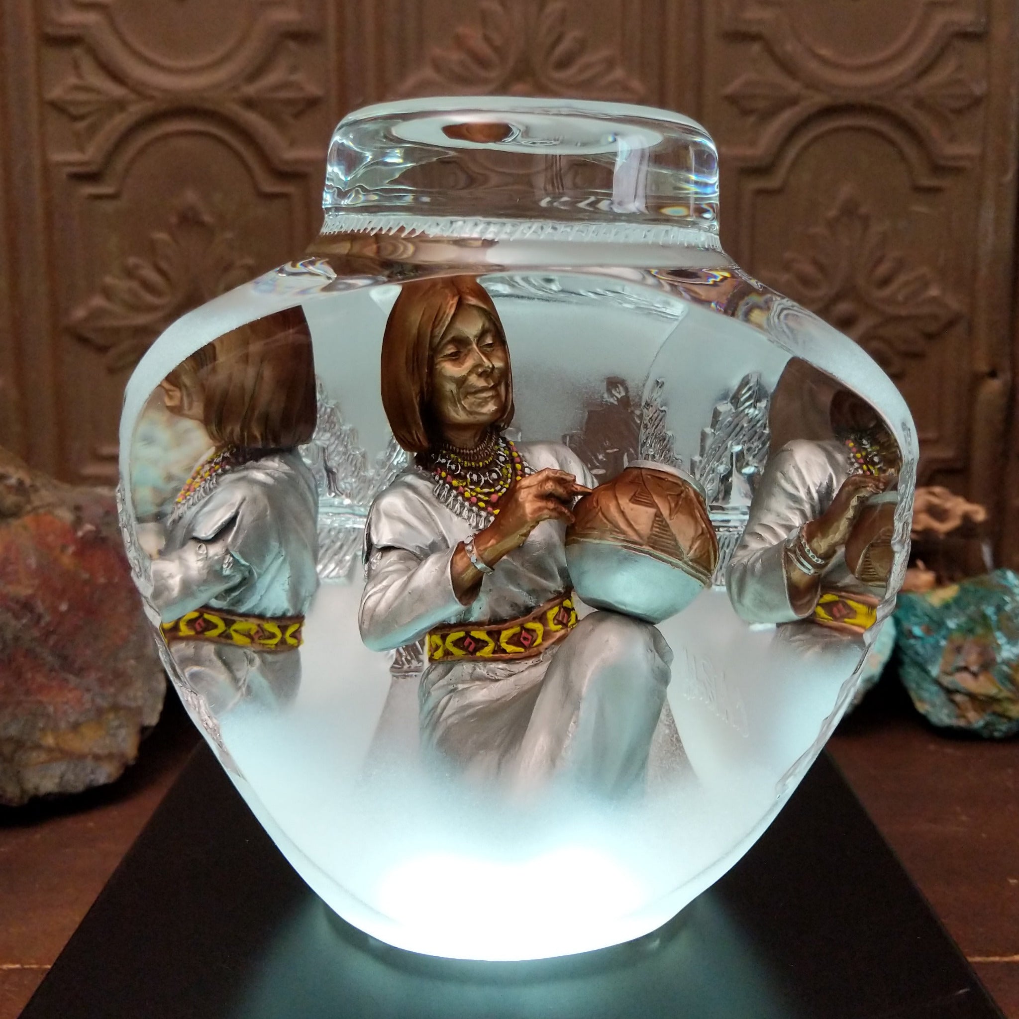 An Indigenous woman decorating a traditional pottery vessel, enclosed in a clear Lucite sculpture that creates mirrored reflections. The figure wears a silver-toned dress with colorful beaded jewelry; lower light display.