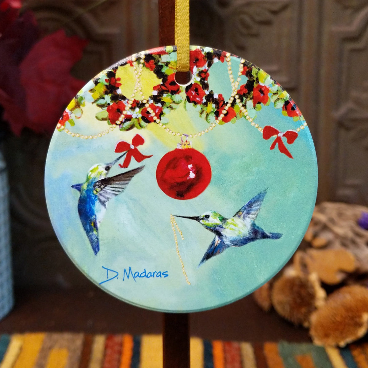 A round Christmas ornament featuring two hummingbirds, a red bauble, and a holiday garland, signed D. Madoras, hanging on a stand.