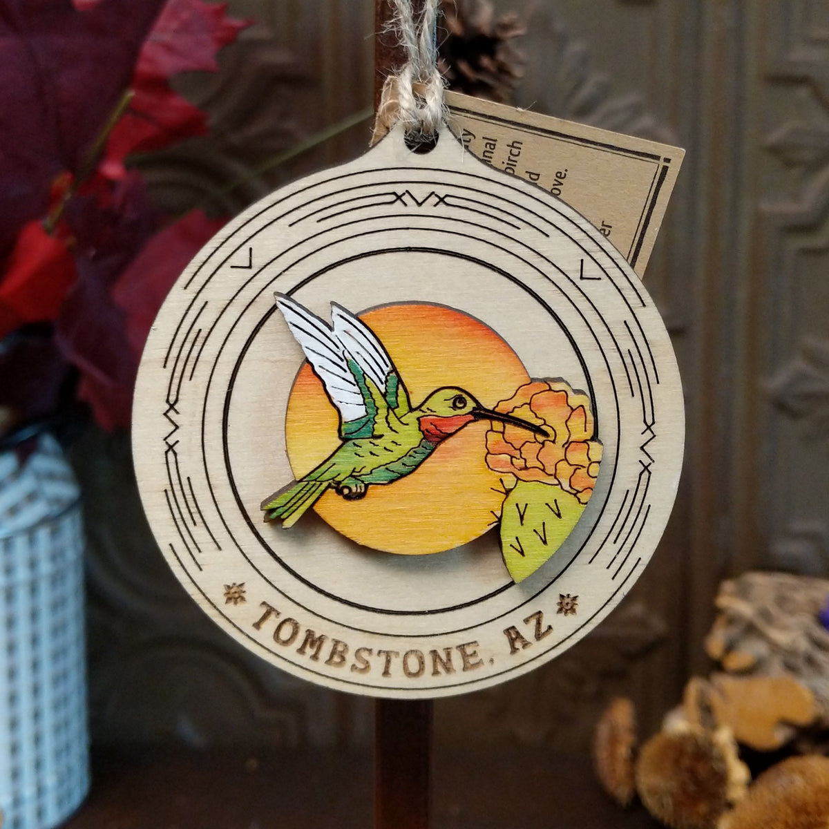 Hummingbird Sunset Wood Ornament featuring a colorful green hummingbird, orange desert flowers, sunset background, engraved circular wooden frame, “Tombstone, AZ” lettering, and natural twine hanging loop.