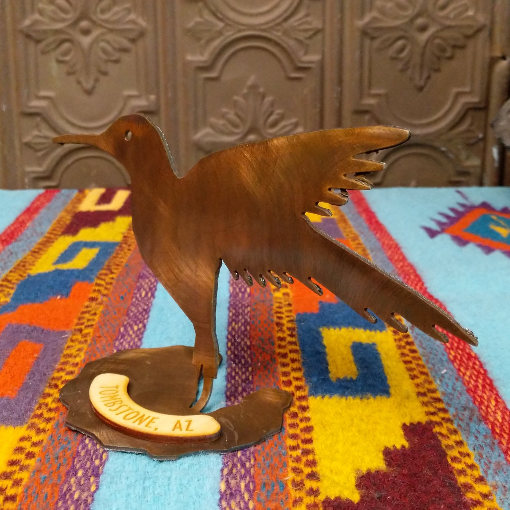 Handcrafted Crooks Design metal hummingbird tabletop sculpture with outstretched wings, detailed feather cutouts, a warm burnished finish, and a decorative base marked Tombstone, AZ; side view.