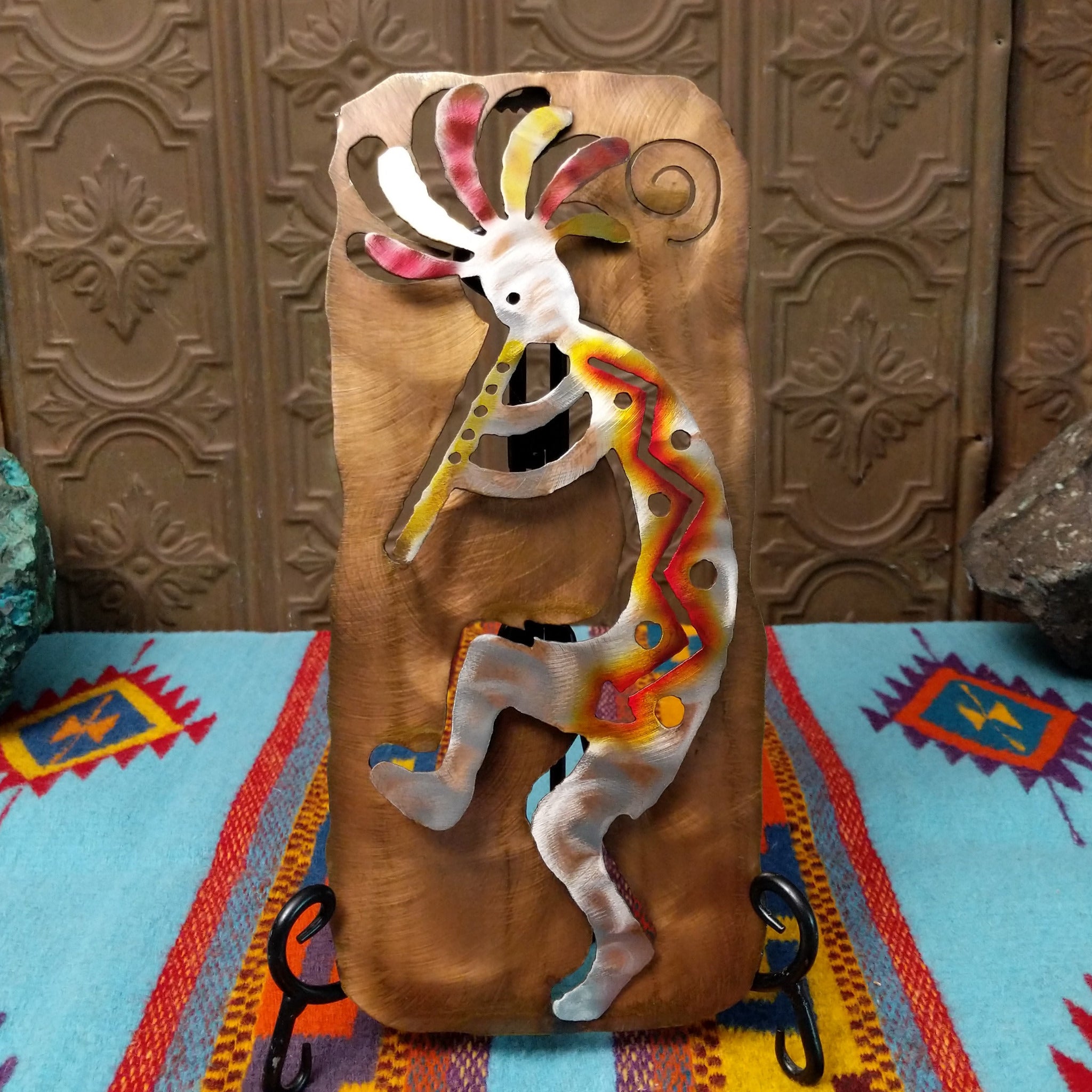 This Crooks Design metal Kokopelli features a brushed silver-toned flute player with red, yellow, and orange accents, a colorful feathered headdress, and a warm textured metal background.
