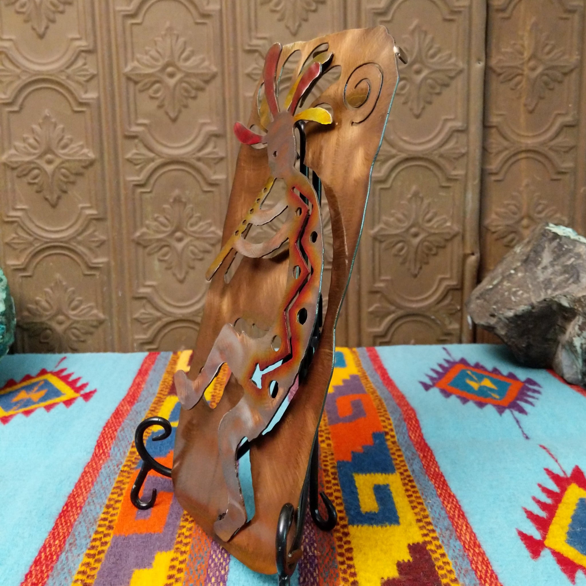 This Crooks Design metal Kokopelli features a brushed silver-toned flute player with red, yellow, and orange accents, a colorful feathered headdress, and a warm textured metal background; side view.