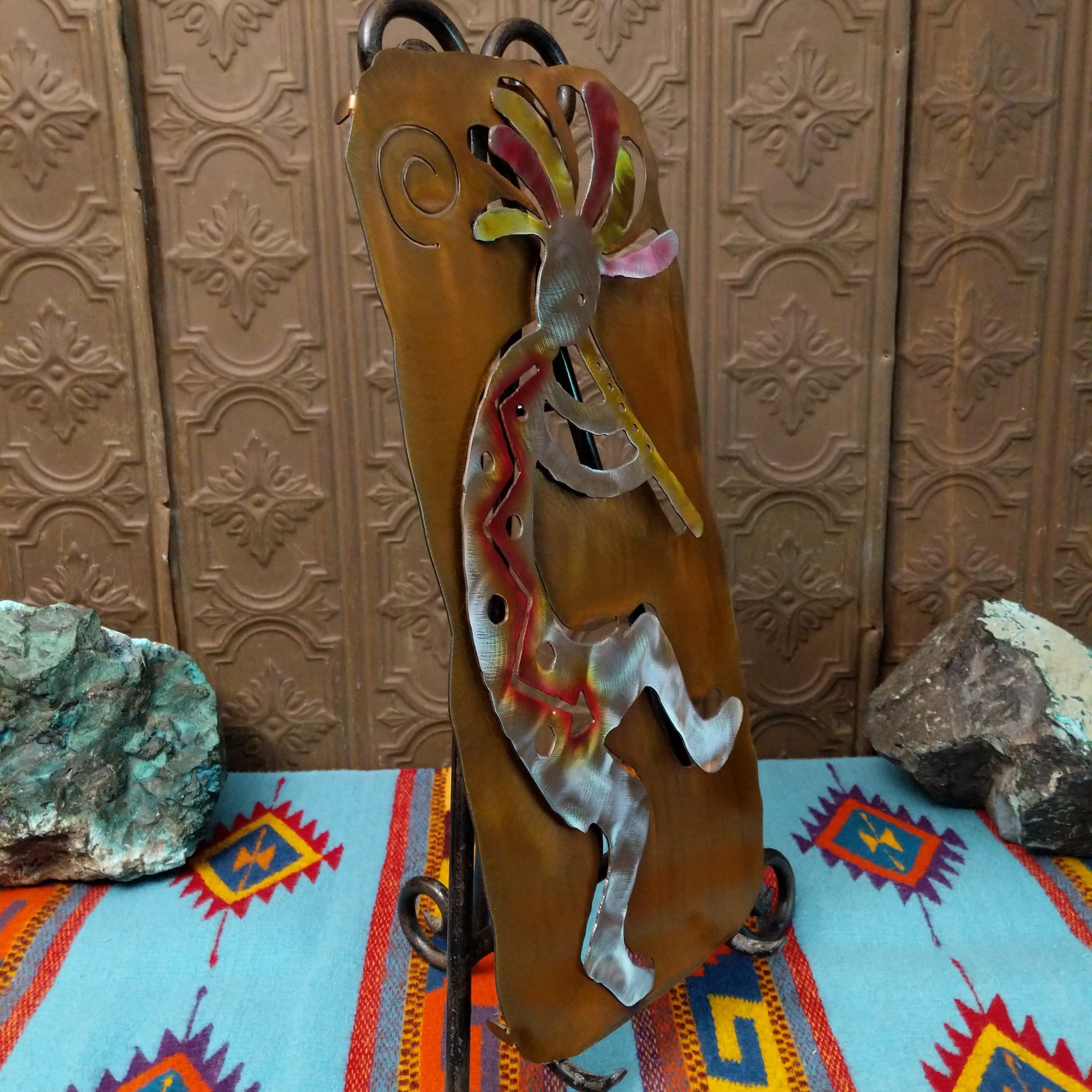Crooks Design metal Kokopelli featuring a brushed silver-toned figure playing a flute, colorful red, yellow, pink, and green accents, and a warm golden-brown metal background; side view.