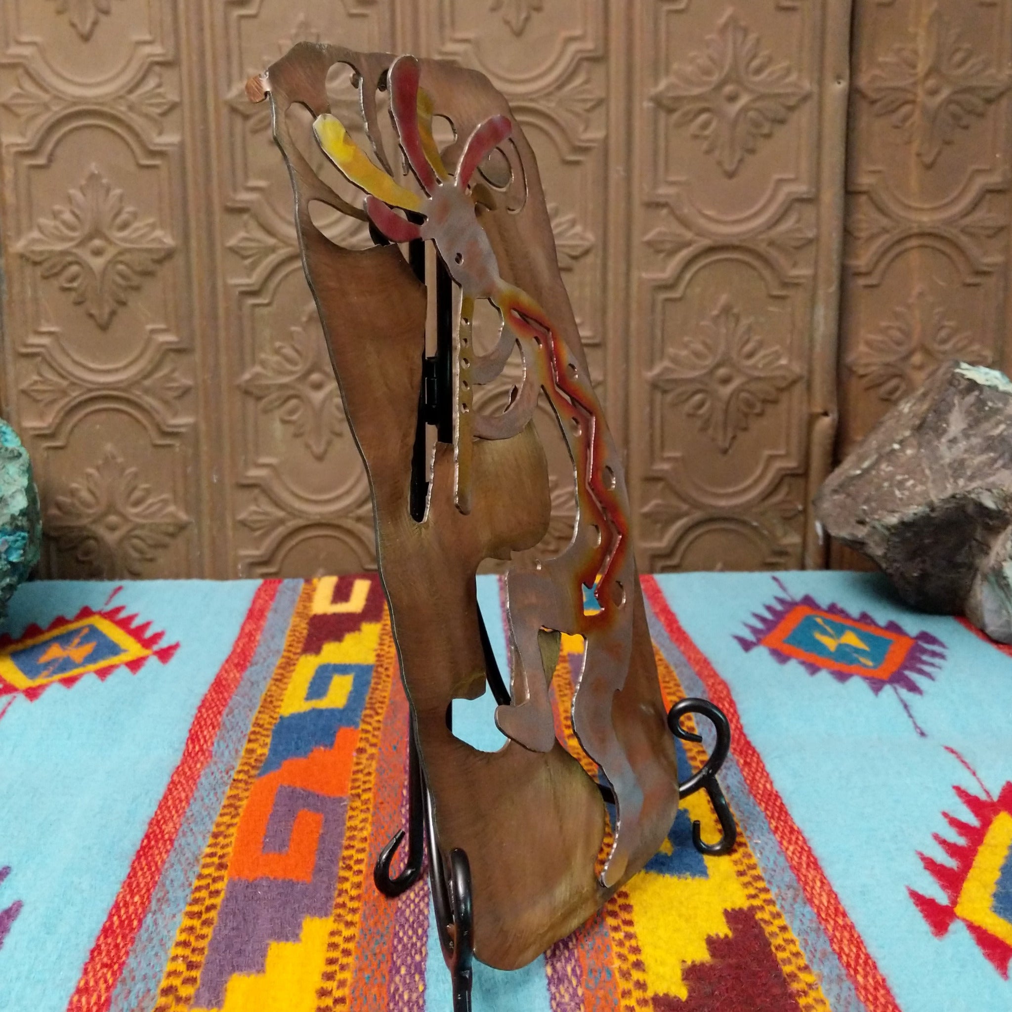 This Crooks Design metal Kokopelli features a brushed silver-toned flute player with red, yellow, and orange accents, a colorful feathered headdress, and a warm textured metal background; side view.