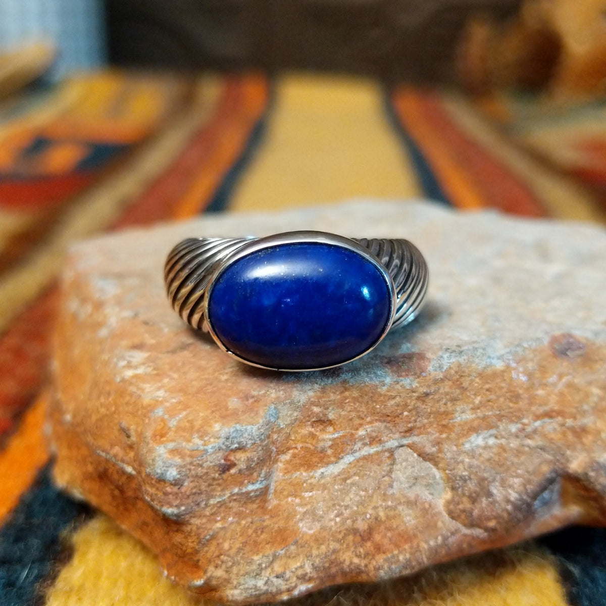 Front view of a silver ring by Elgin Tom featuring a horizontal oval blue lapis lazuli cabochon and a wide band with diagonal ribbed detailing.