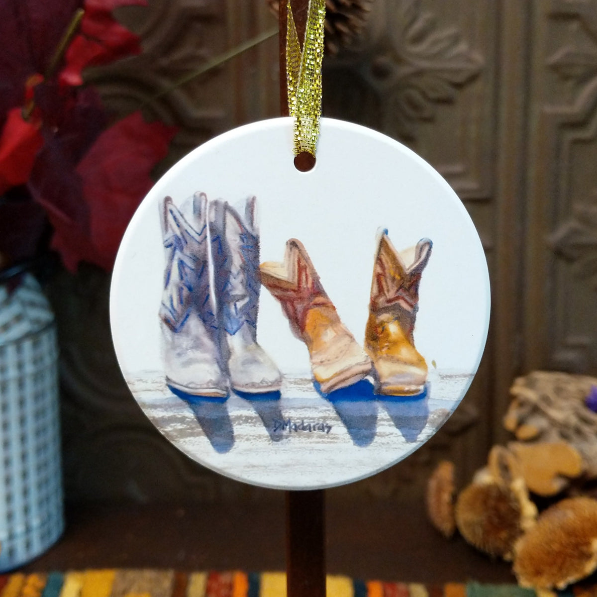 Round ceramic Christmas ornament featuring a painted illustration of three cowboy boots in white, tan, and brown, hanging by a gold ribbon against a rustic holiday background.