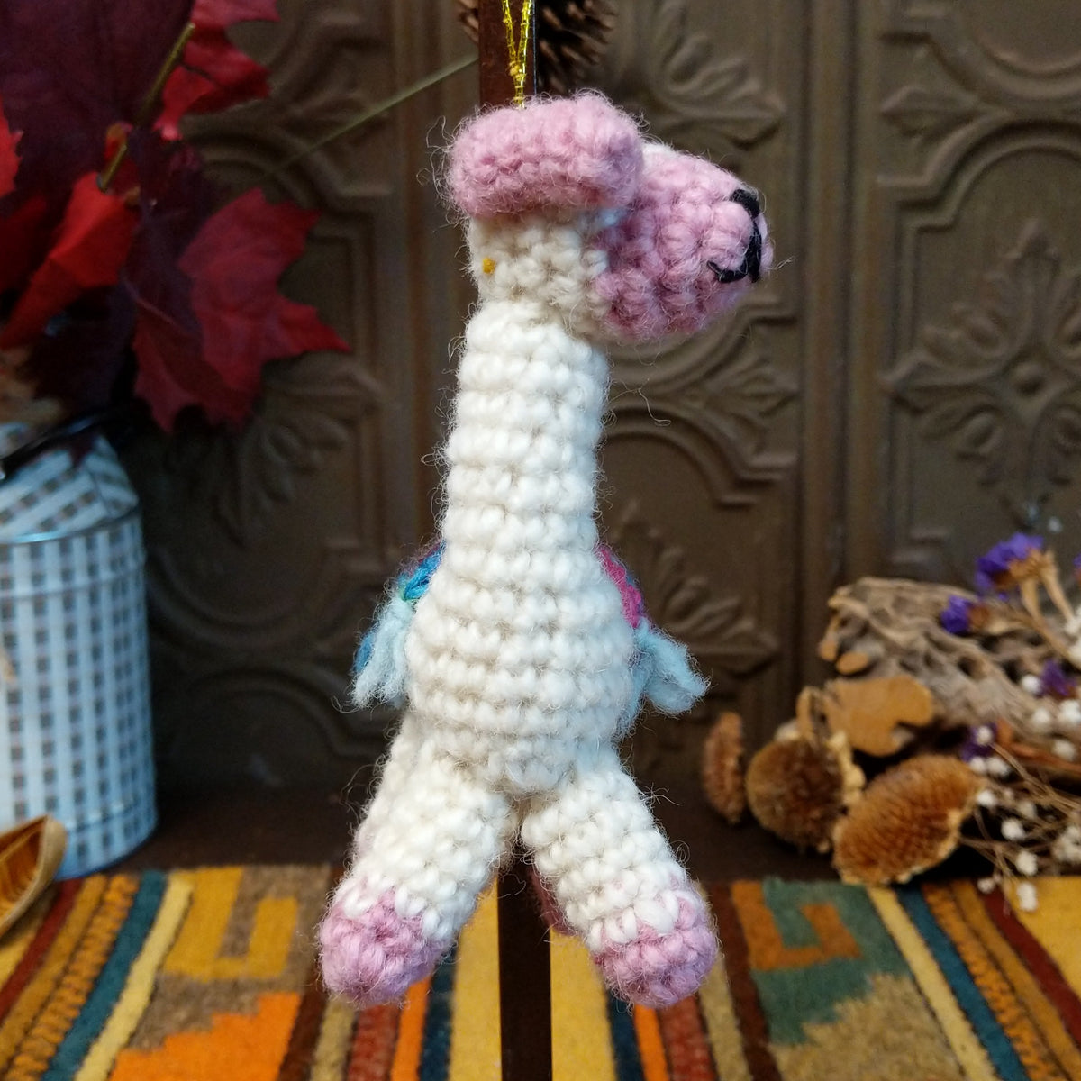 This charming Llama Wool Ornament features a white crocheted body, pastel pink face, ears, hooves, and curled tail, a colorful Southwestern-inspired saddle blanket, black embroidered facial details, and a gold hanging cord.
