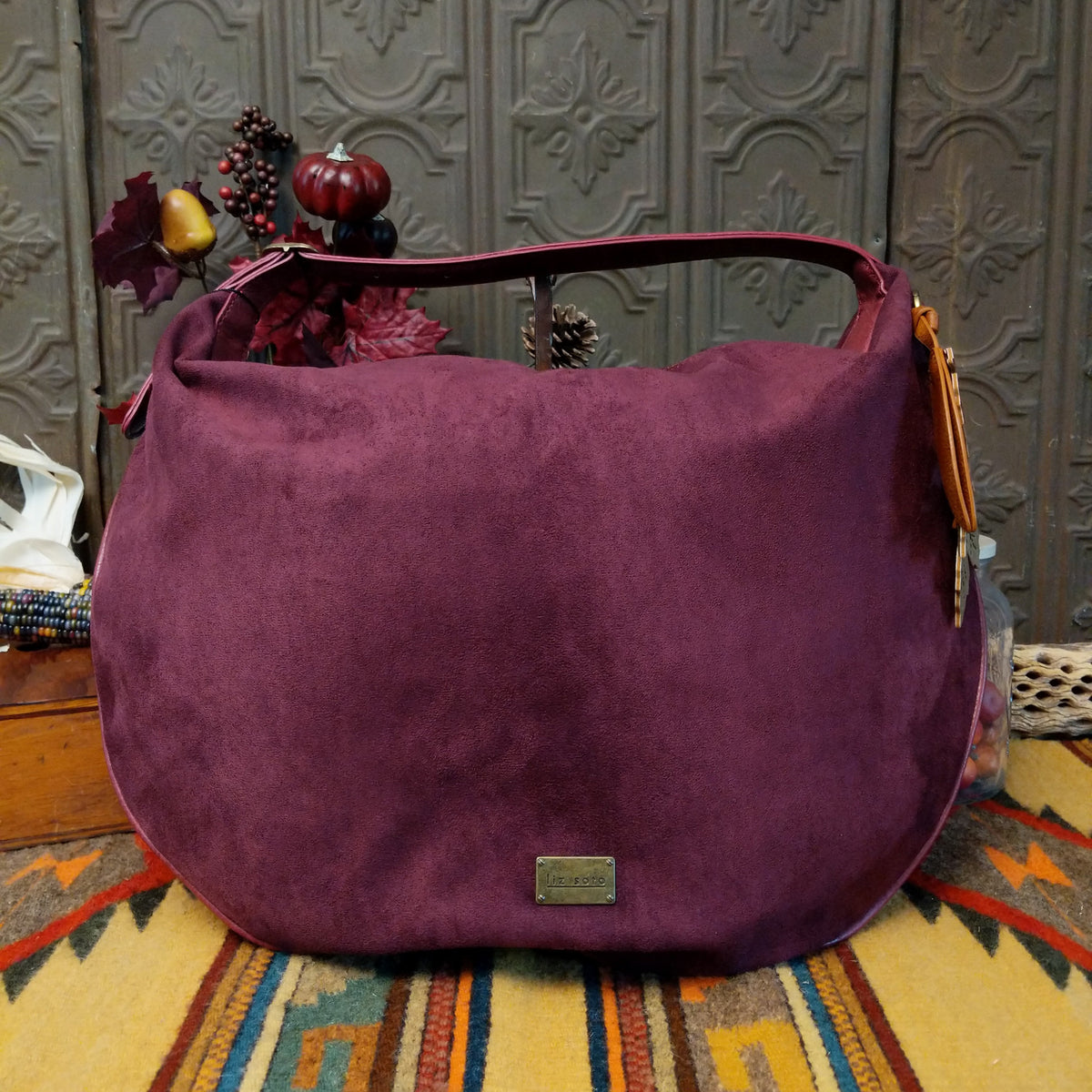 Lulu Handbag by Liz Soto featuring a spacious hobo silhouette, soft suede-like exterior, long shoulder strap, side buckle detailing, top zip closure, and logo plaque.
