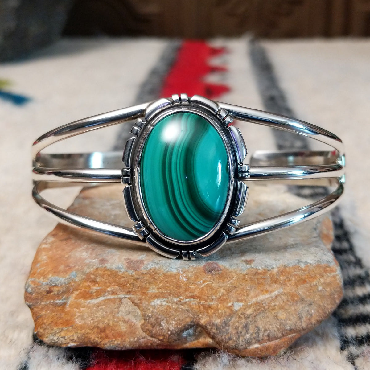 Silver cuff by Amory Begay featuring a large oval malachite stone with sweeping bands of dark and vivid green, surrounded by geometric silver accents and split silver bands.