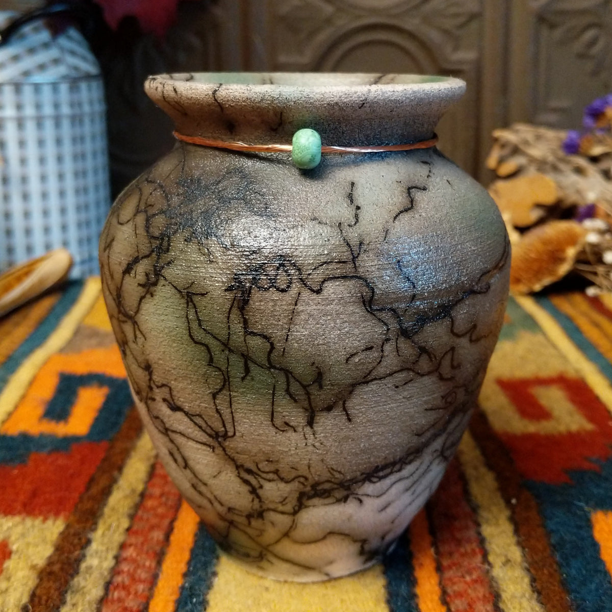 This charming Mini Mottled Green Horsehair Pottery by Roger Calhoun features a rounded vase with a flared black rim, earthy tones, soft green accents, intricate black horsehair patterns, and a copper wire with a green bead.