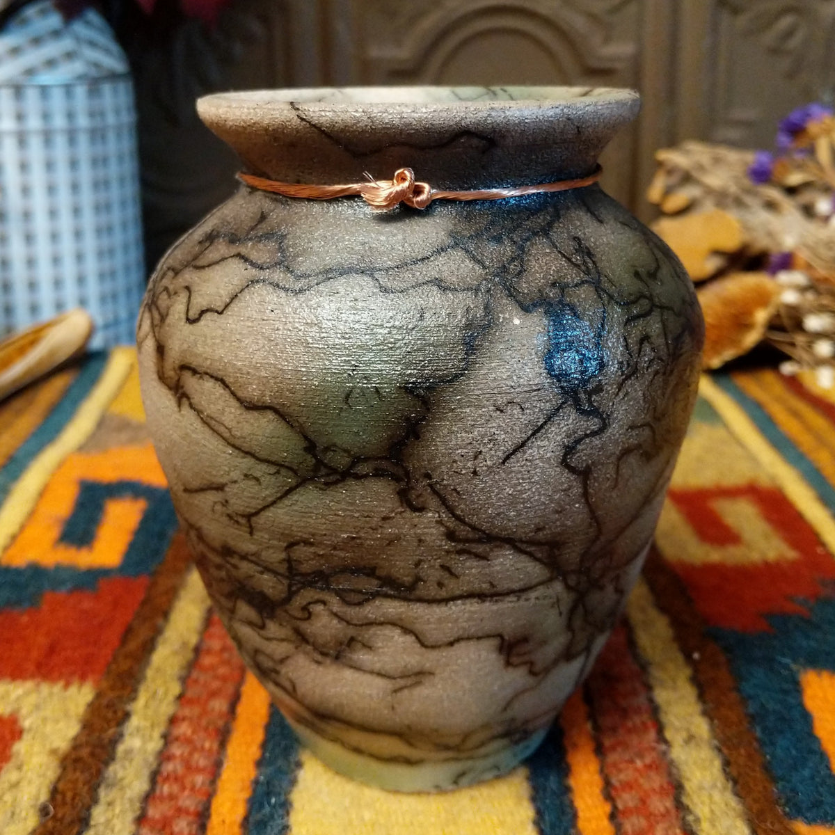 This charming Mini Mottled Green Horsehair Pottery by Roger Calhoun features a rounded vase with a flared black rim, earthy tones, soft green accents, intricate black horsehair patterns, and a copper wire with a green bead.