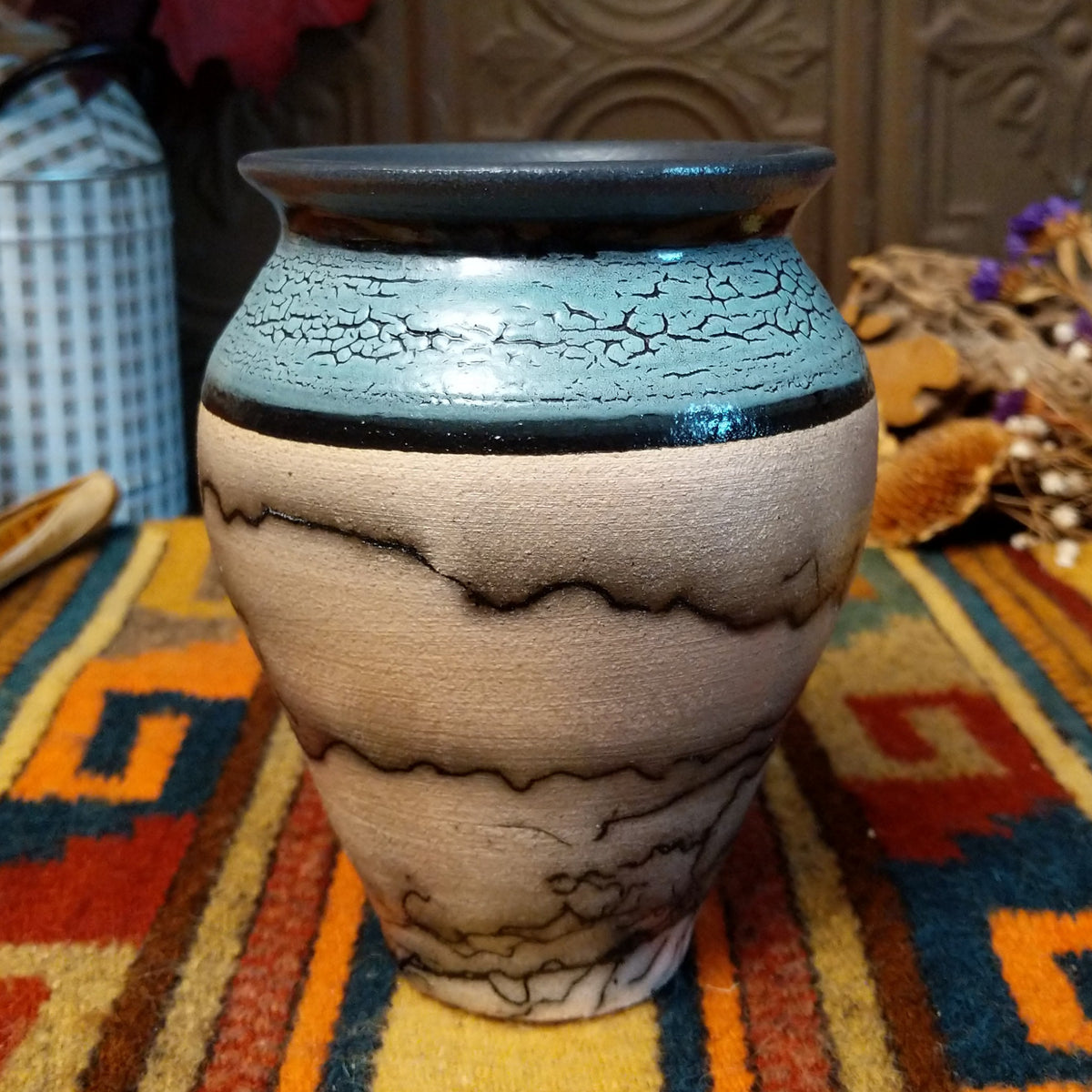 Mini horsehair pottery vase by Roger Calhoun with a flared dark rim, crackled turquoise-blue shoulder, and black horsehair markings concentrated near the base of its cream-colored body.