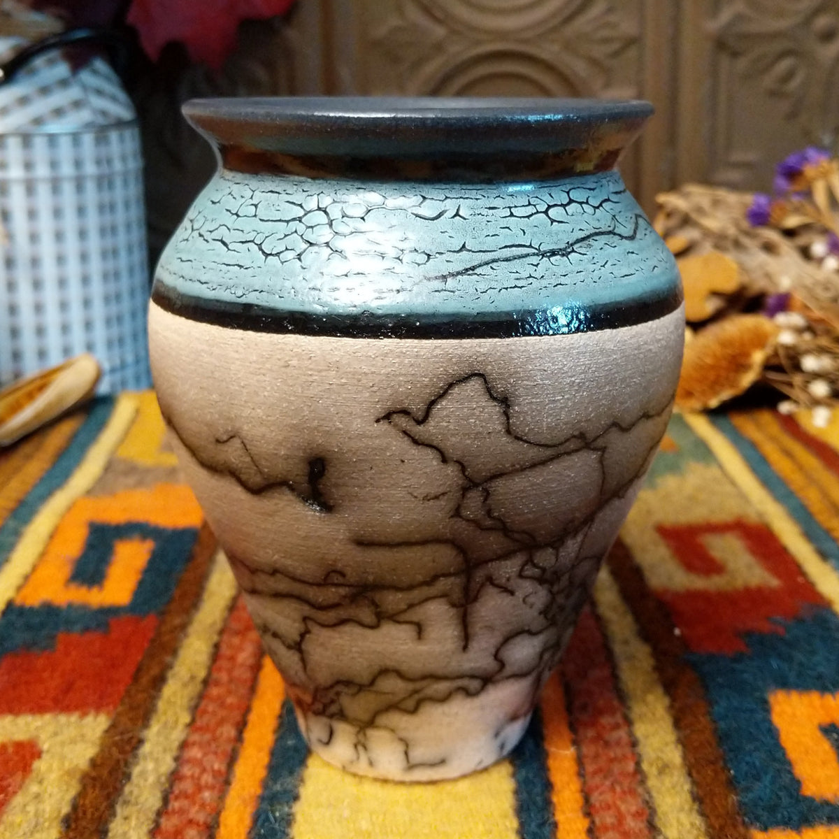 Mini horsehair pottery vase by Roger Calhoun with a flared dark rim, crackled turquoise-blue shoulder, and black horsehair markings concentrated near the base of its cream-colored body.