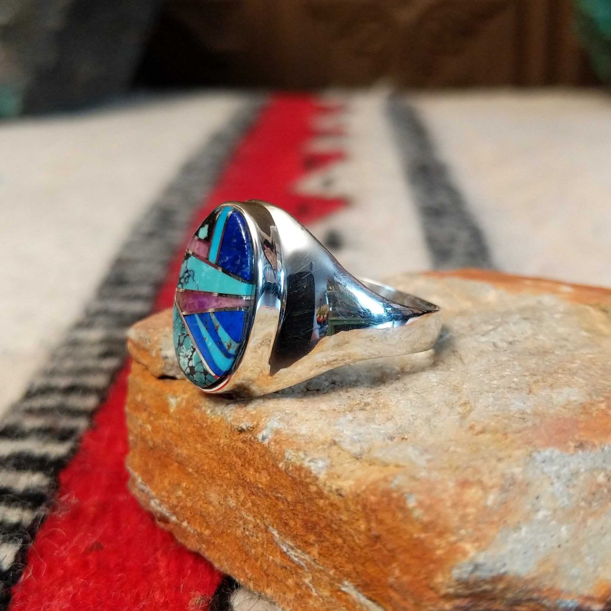Multistone inlay ring by P.D. featuring an oval geometric design with turquoise, blue, purple, and black stones set in a polished silver band; side view.