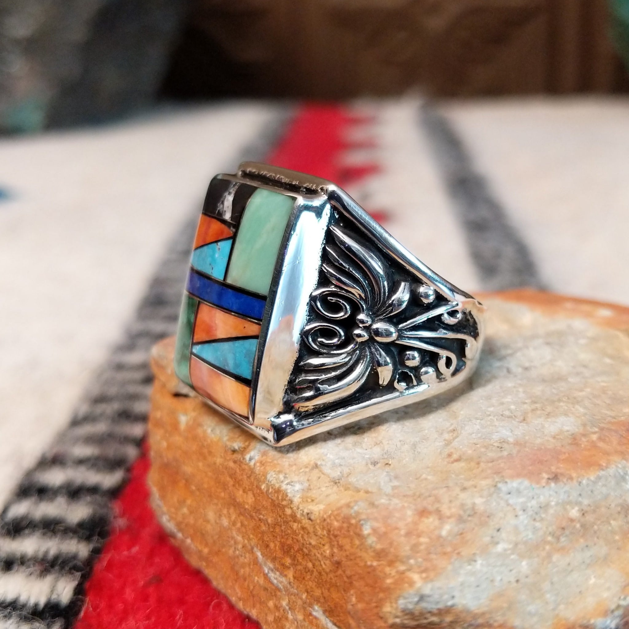 Multistone inlay ring by Terry Wood featuring a rectangular geometric design with turquoise, blue, green, orange, peach, and black stones set in silver with detailed scrollwork along the sides; side view.