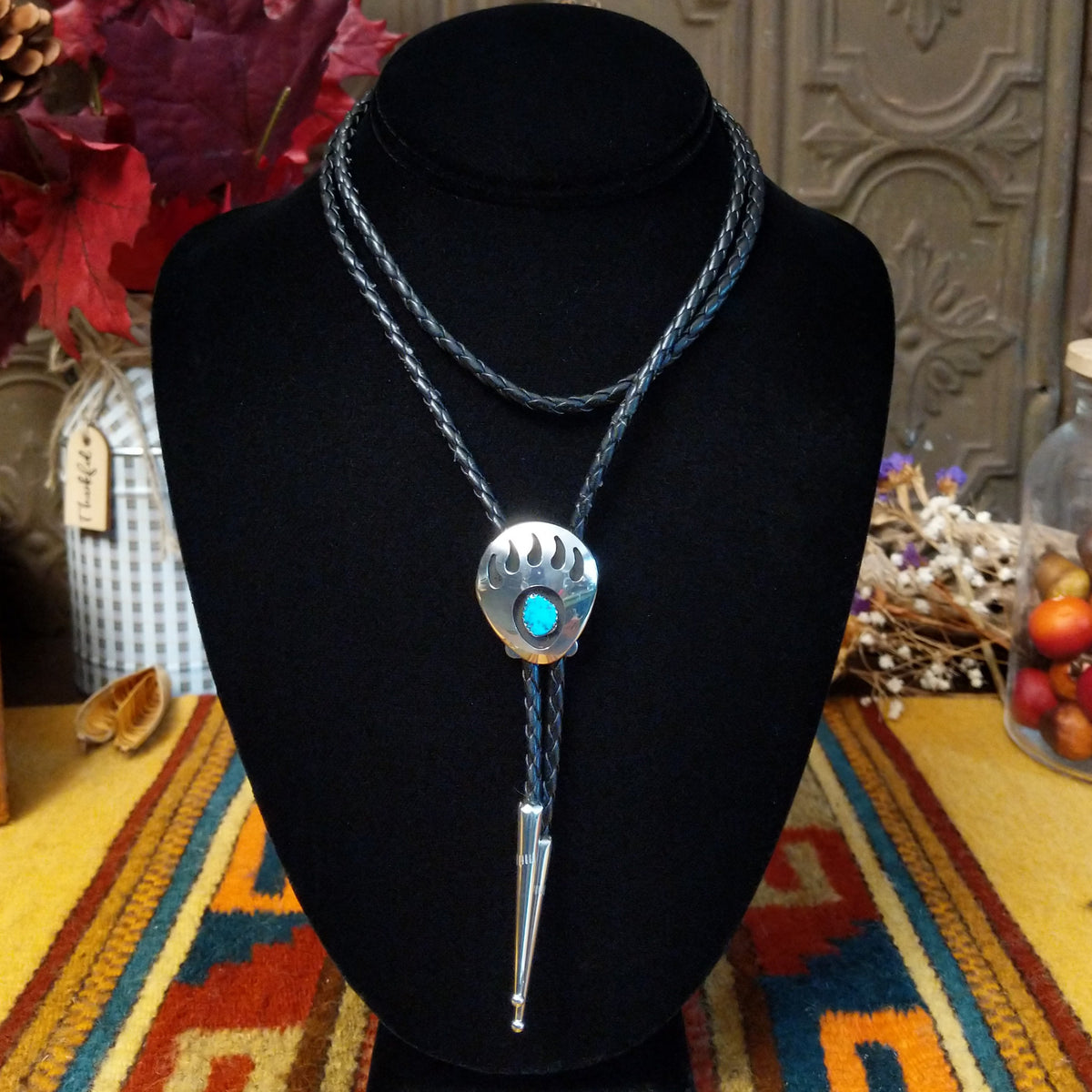 A Sterling Silver bolo tie necklace with a turquoise stone and a paw print pendant, displayed on a black velvet mannequin bust. The necklace features braided blue cords and is set against a rustic background with autumn decorations and a patterned textile.