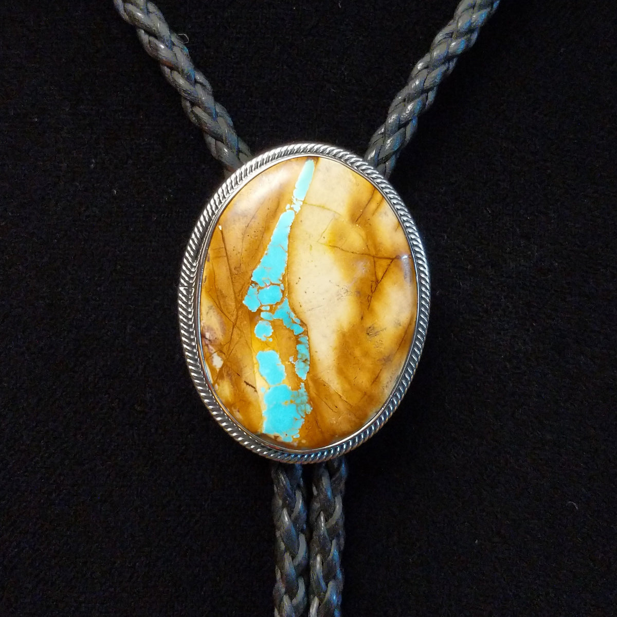 A Sterling Silver bolo tie necklace featuring an oval Boulder Turquoise pendant, suspended on a braided cord against a black background.