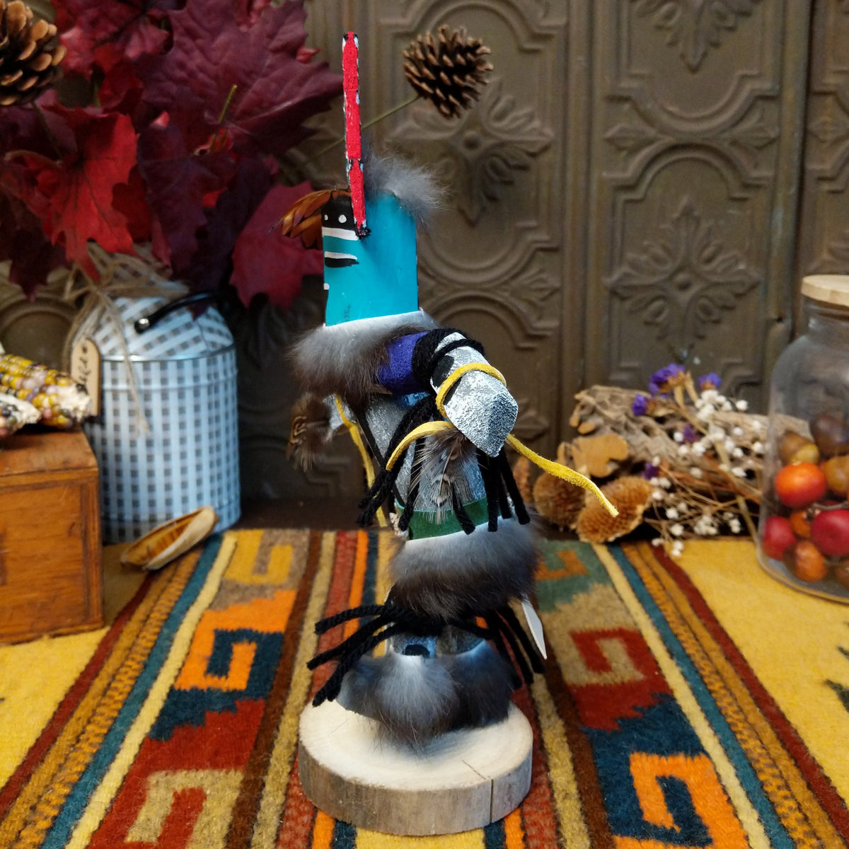 This vibrant Navajo Hemis Kachina by Debbie James features a hand-painted red and turquoise mask, stepped headdress, natural feathers, soft fur accents, leather ties, and a circular wooden display base; side view.