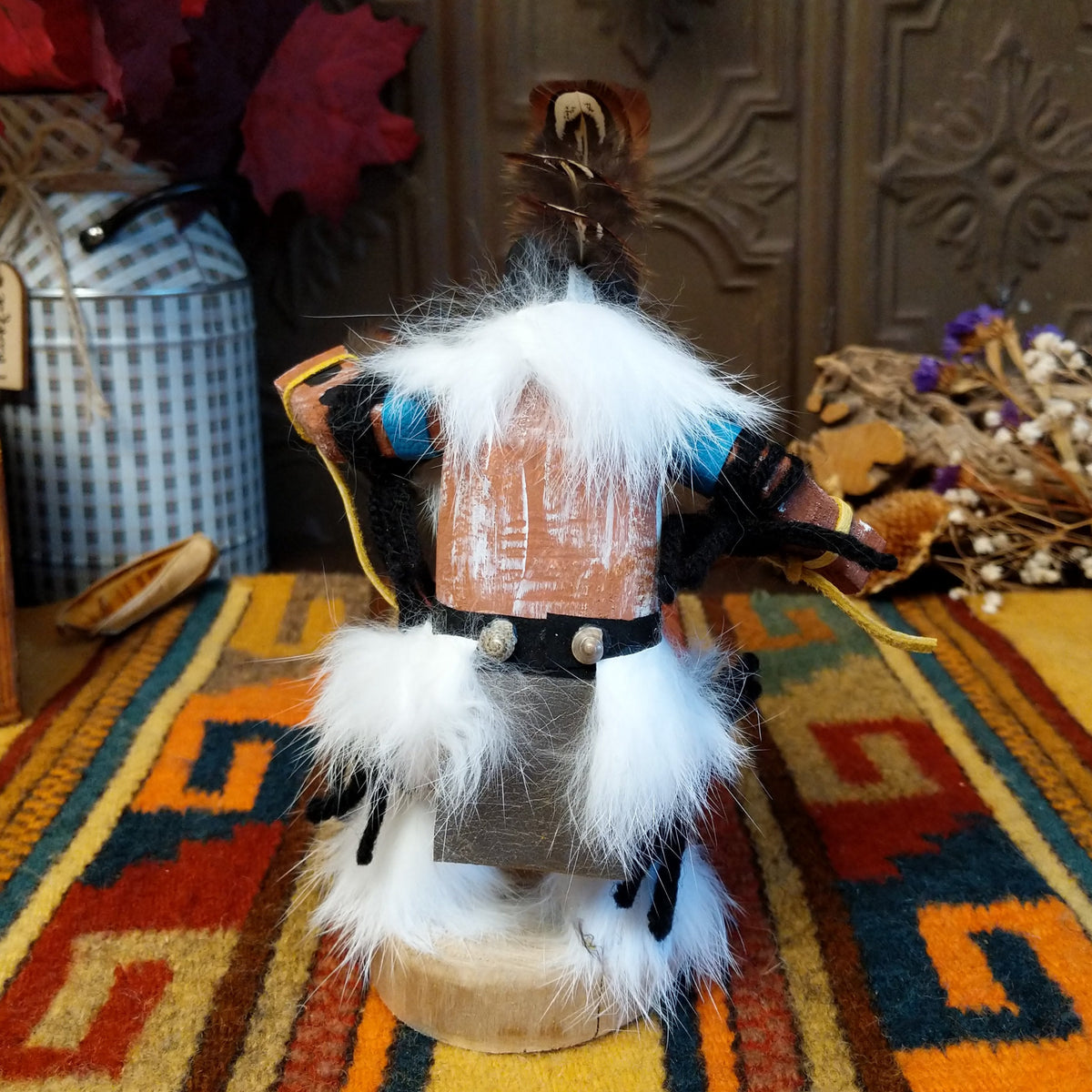 This distinctive Navajo Kokopelli Kachina by Debbie James features a hand-painted wooden figure with a tall feather headdress, black face, white fur accents, natural feathers, leather ties, and a wooden display base; back view.