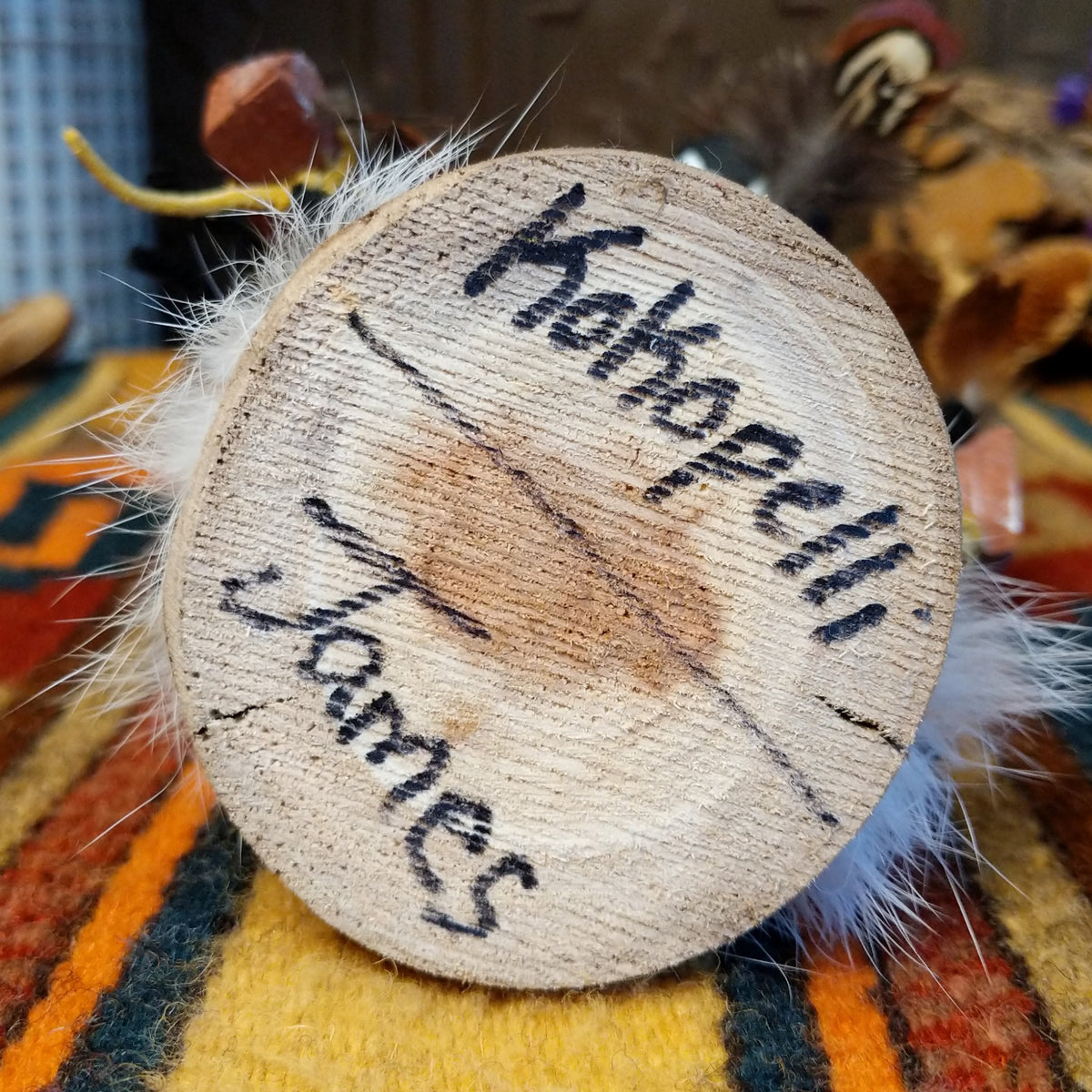 This distinctive Navajo Kokopelli Kachina by Debbie James features a hand-painted wooden figure with a tall feather headdress, black face, white fur accents, natural feathers, leather ties, and a wooden display base; artisan and product name on bottom.
