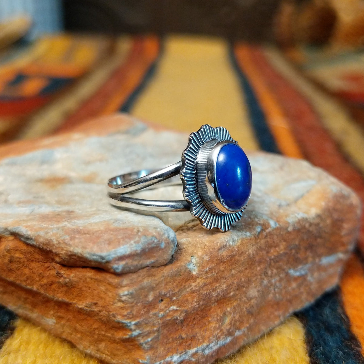 Angled side view of a silver ring featuring a raised oval blue lapis lazuli cabochon, radiating stamped silverwork, and a polished split-shank band.