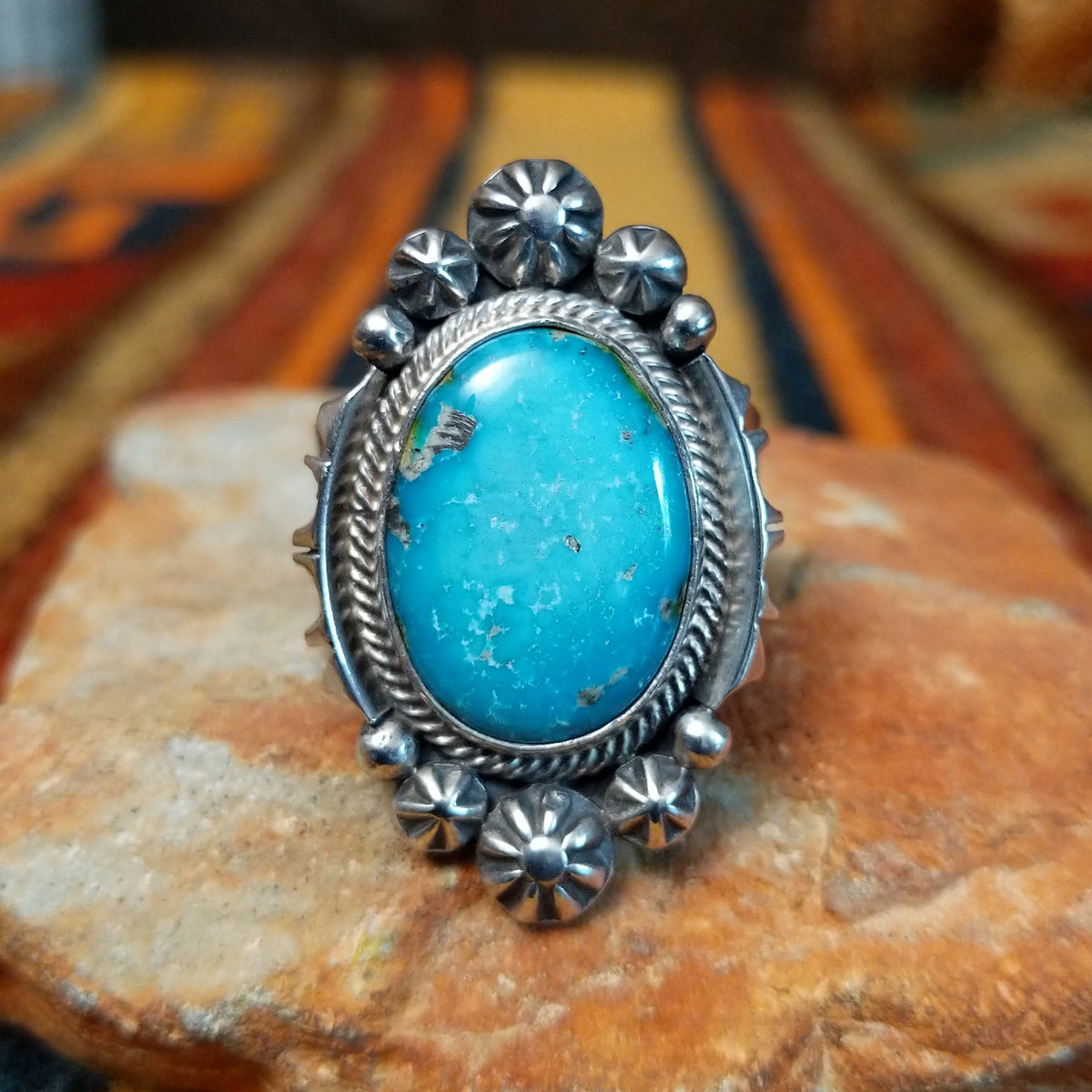 This elegant Navajo Ornate Sonoran Gold Turquoise Ring features a large oval blue turquoise stone surrounded by a silver rope border, polished silver beads, and decorative stamped silver blossoms.