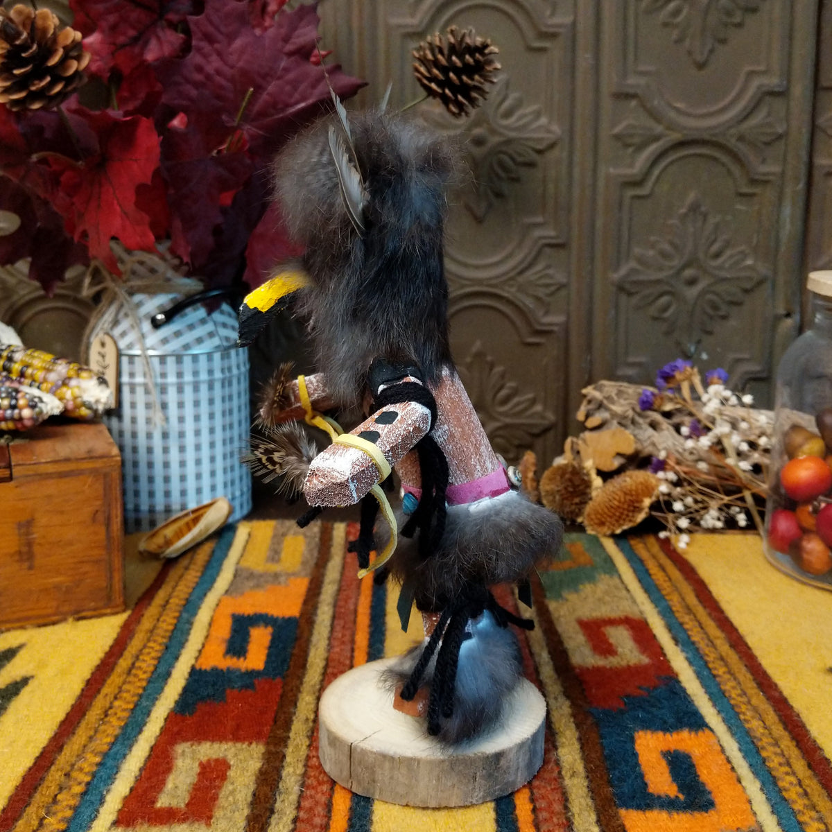 This unique Navajo Owl Kachina by Debbie James features a hand-painted wooden figure with a yellow and black owl beak, dark fur headdress, natural feathers, leather ties, decorative clothing, and a wooden display base; side view.
