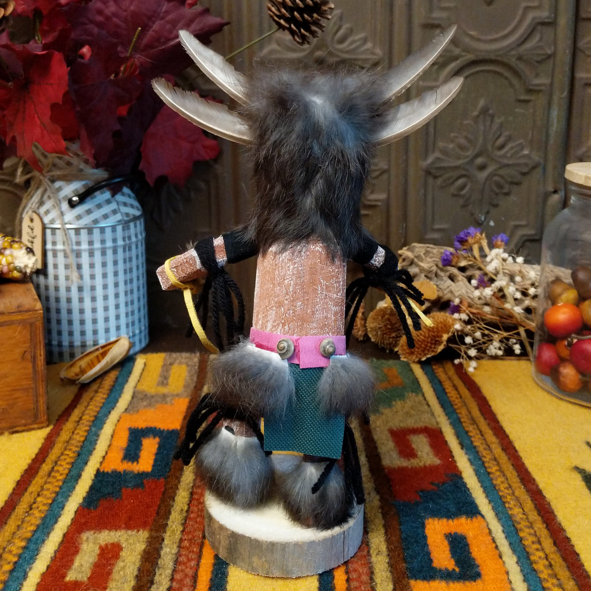 This unique Navajo Owl Kachina by Debbie James features a hand-painted wooden figure with a yellow and black owl beak, dark fur headdress, natural feathers, leather ties, decorative clothing, and a wooden display base; back view.