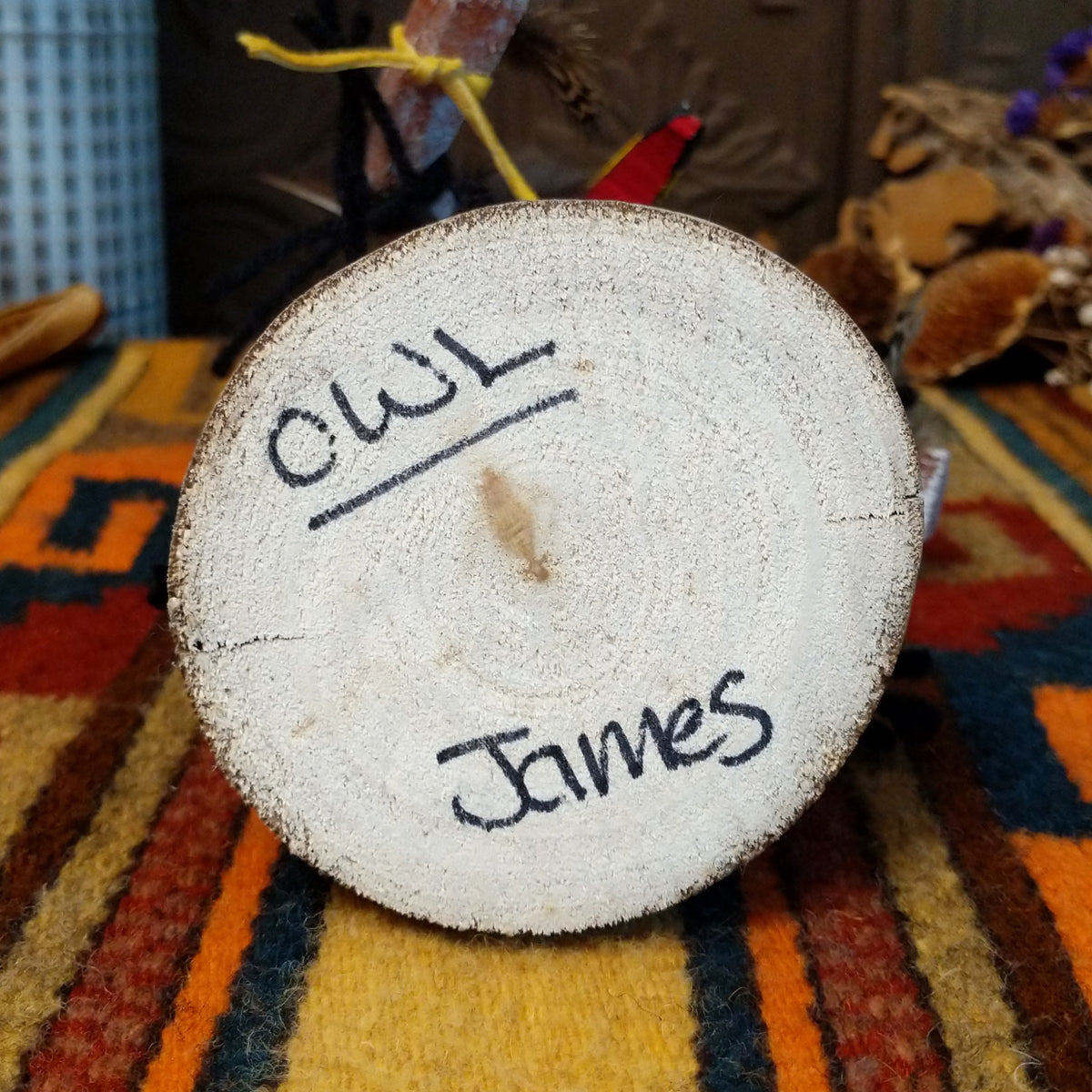 This unique Navajo Owl Kachina by Debbie James features a hand-painted wooden figure with a yellow and black owl beak, dark fur headdress, natural feathers, leather ties, decorative clothing, and a wooden display base; artisan and product name on bottom.