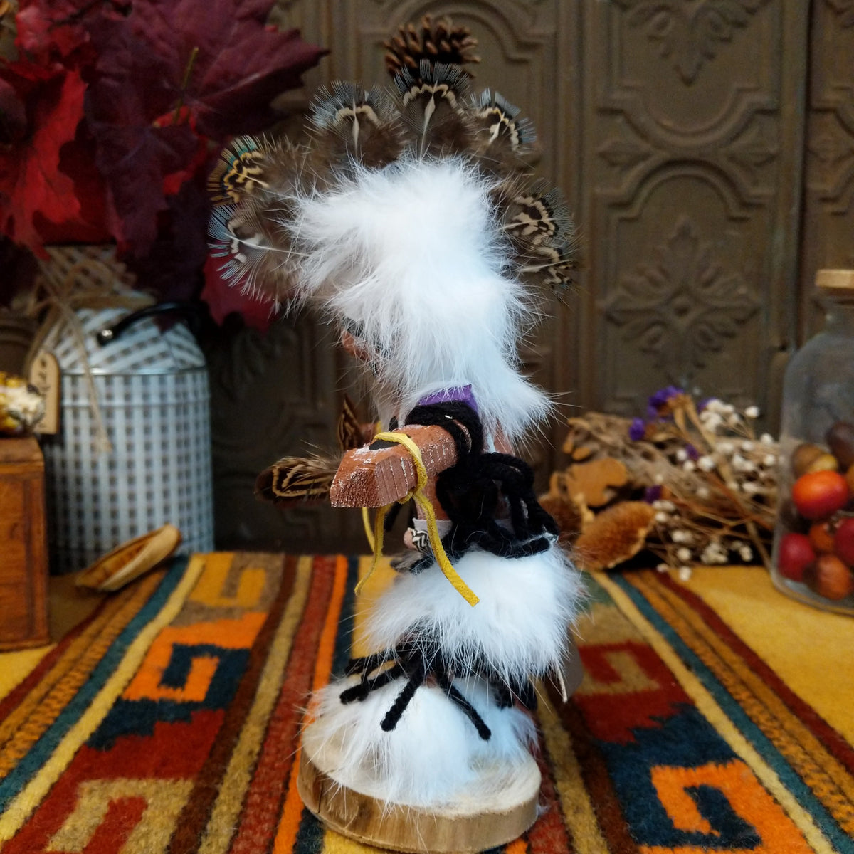 This distinctive Navajo Protection Dancer Kachina by Debbie James features a hand-painted wooden figure with a white feather headdress, natural feathers, leather ties, fur accents, colorful fabric details, and a wooden display base; side view.