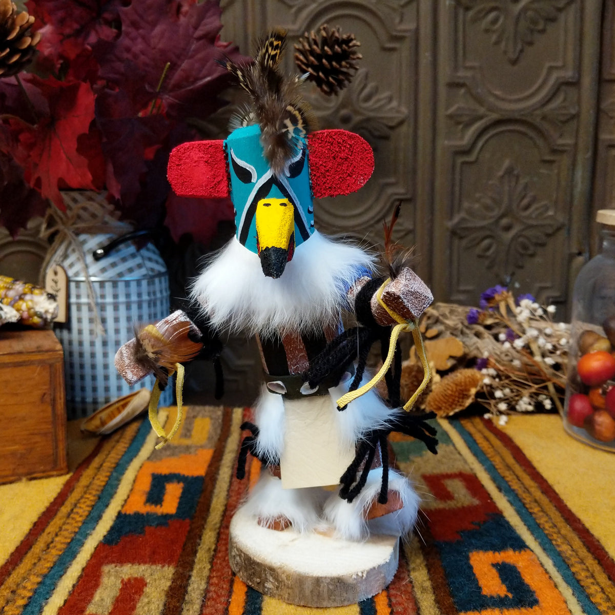 This colorful Navajo Quail Kachina by Debbie James features a turquoise hand-painted bird mask with red accents, a yellow beak, natural feather crest, white fur, leather ties, and a wooden display base.